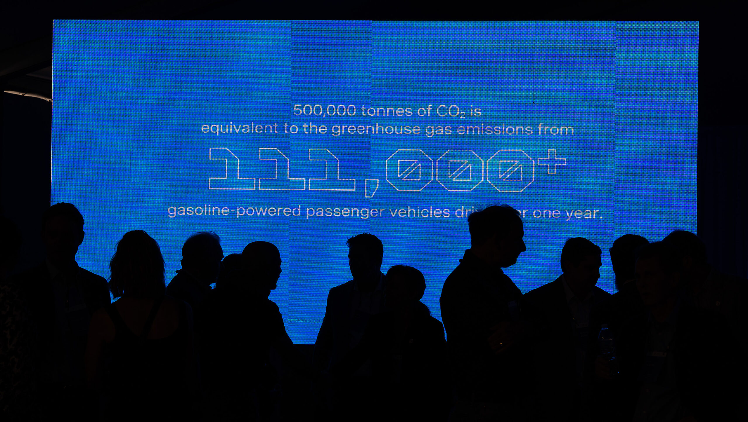 The silhouettes of people standing in front of a large screen that says “500,000 tonnes of CO2 is equivalent to the greenhouse gas emissions of from 111,000+ gasoline-powered passenger vehicles”.
