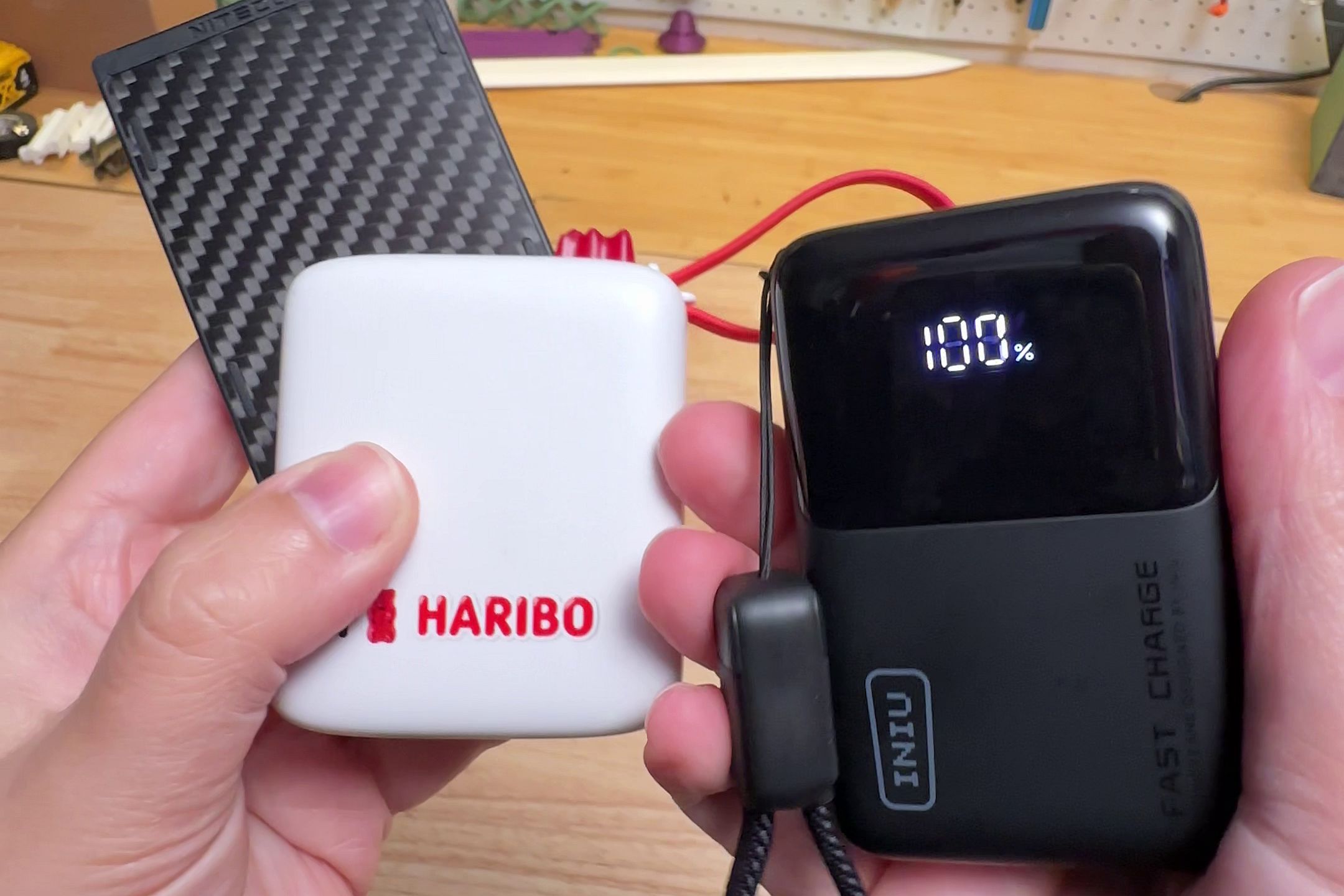 Nitecore, Haribo, and Iniu batteries in my two hands. The Haribo is white with red text, the Nitecore is thin and clad with carbon fiber, and the Iniu is black with a 100% indicator on its tiny display.