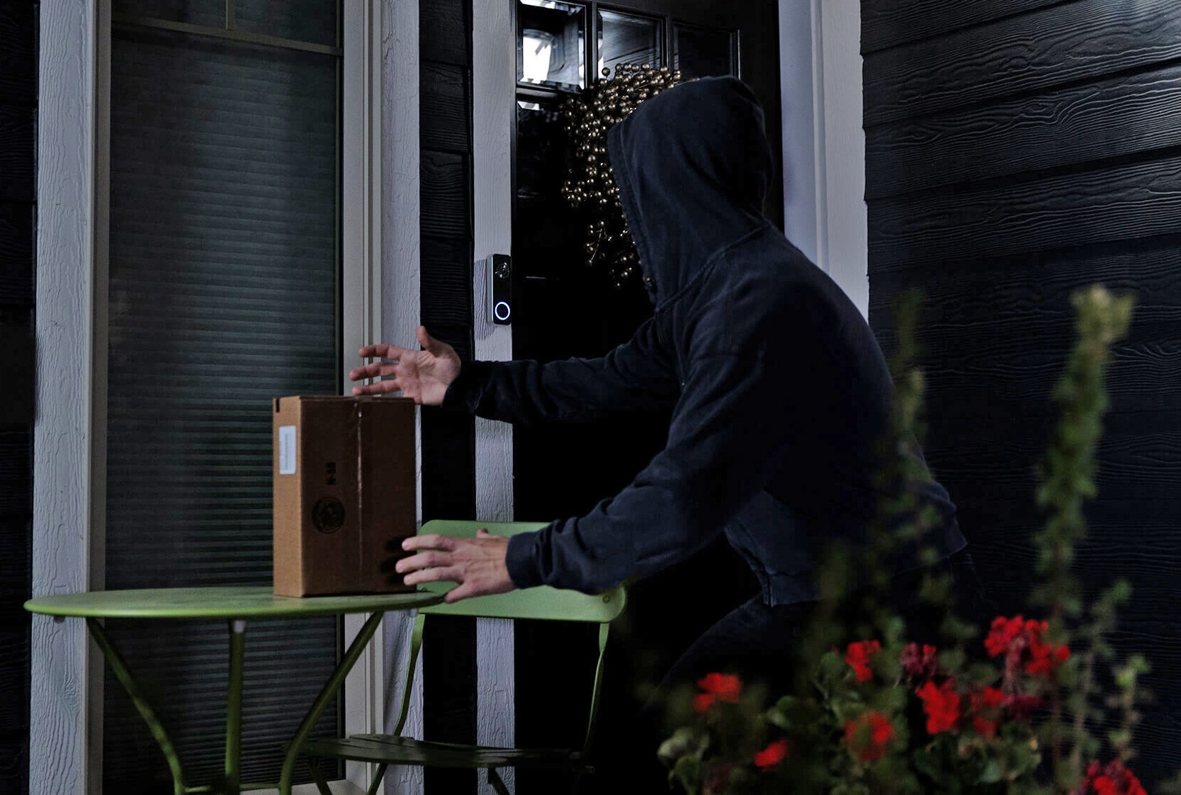 A person wearing a hoodie attempts to grab a boxed package off of a table on a front porch.