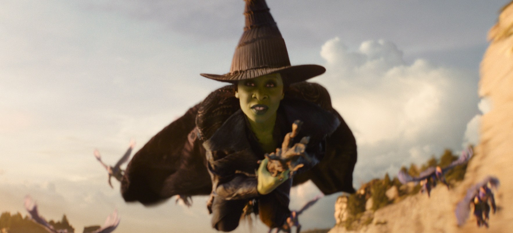 A green-skinned witch wearing a black outfit and a pointy hat while flying on a broomstick over a field.