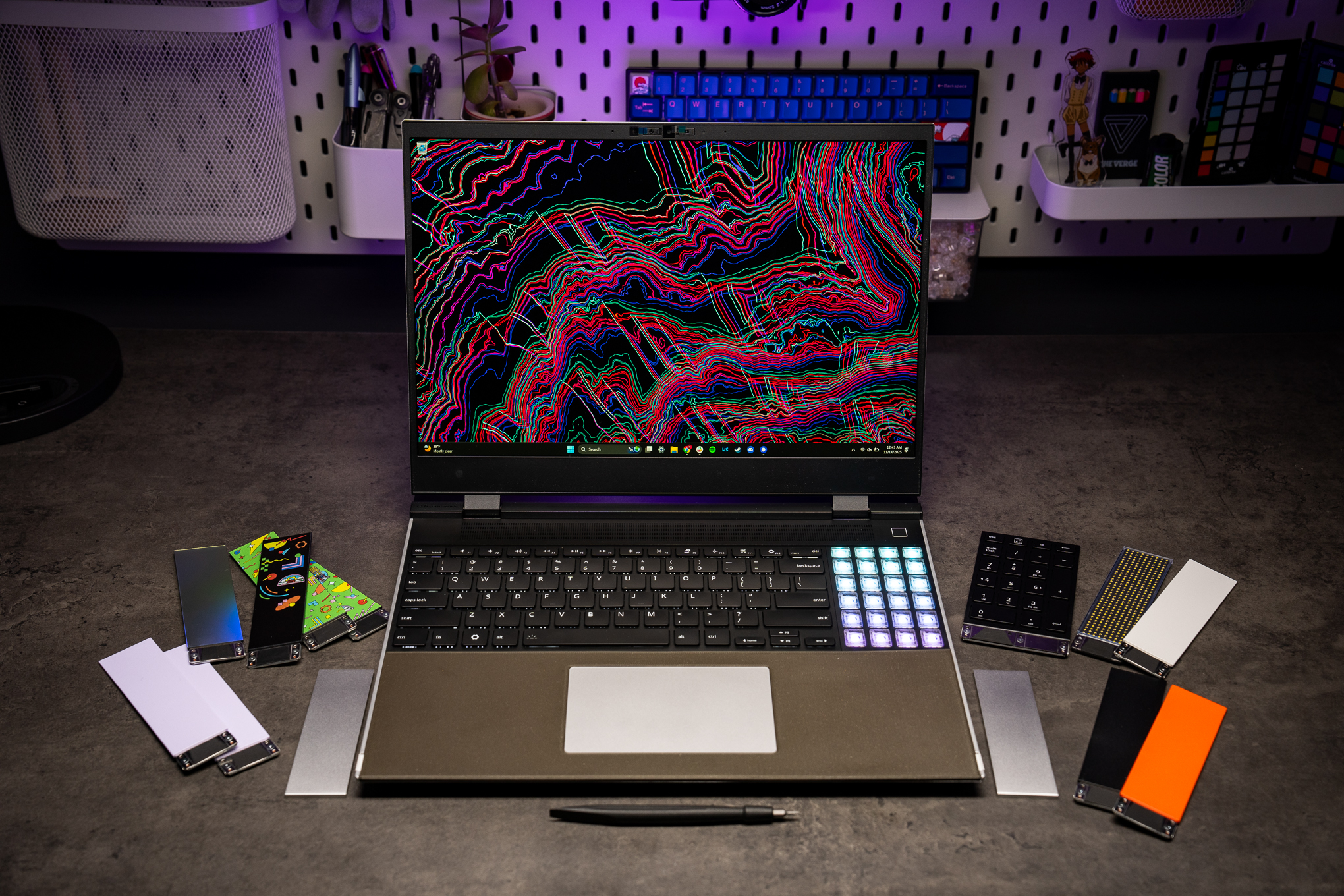 A 2025 Framework Laptop 16 on a desk with keyboard, RGB macro pad, and prototype single-piece trackpad all attached to it. Beside it are a collection of decorative modules that can be attached.