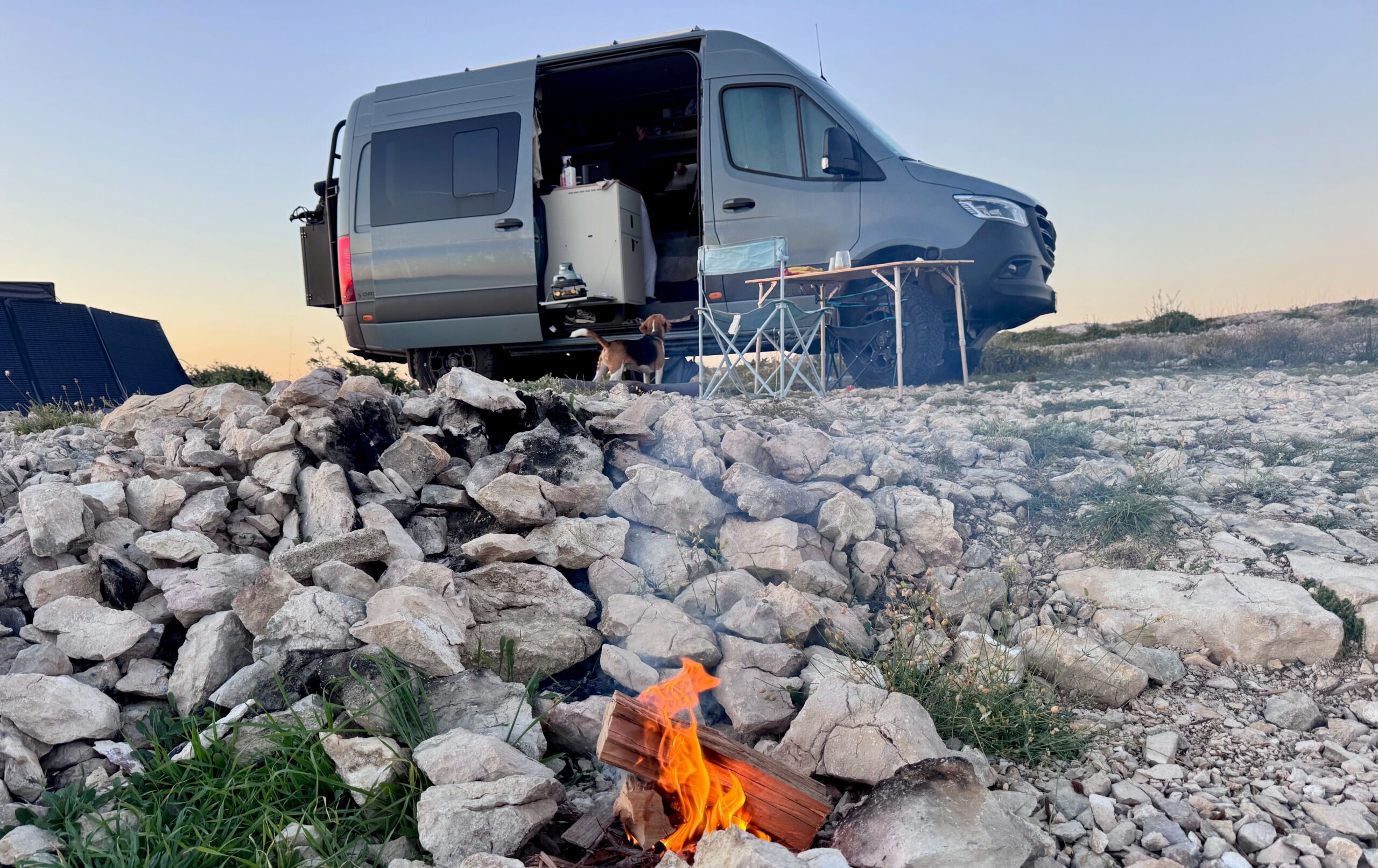 A solar generator with wheels from EcoFlow sits on a gravel road in front of a large off-road tire connected to a lifted 4x4 van with a forest in the background.