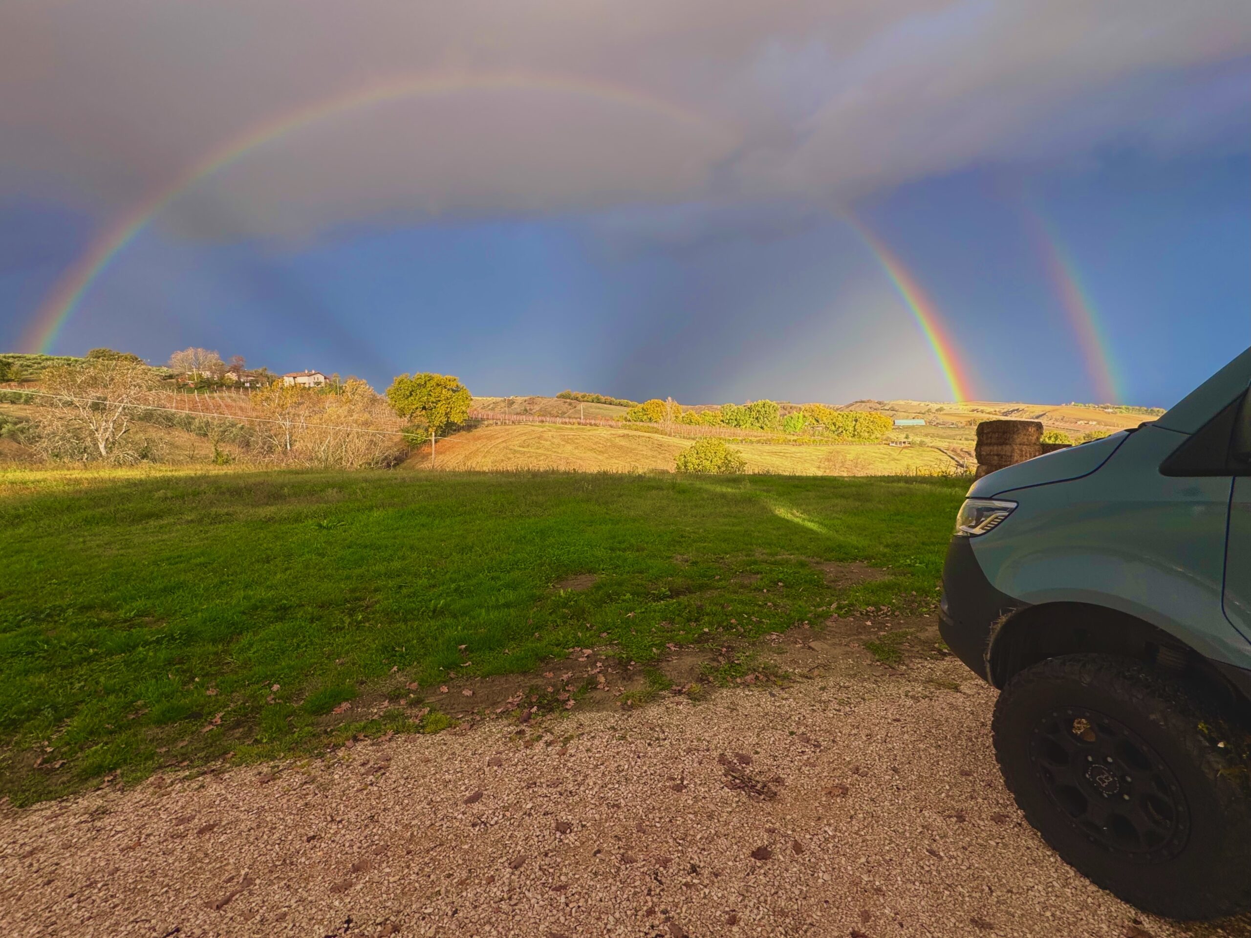 A van is parked on the edge of a vineyard with a dramatic sky and double rainbow in the distance.