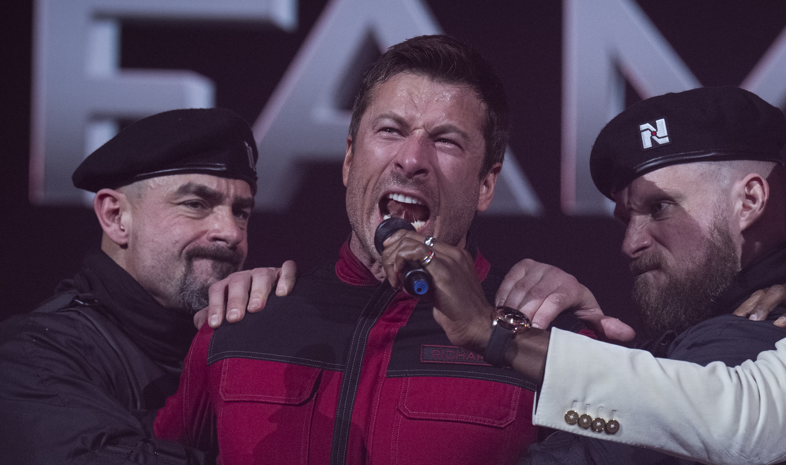 A man in a red and black jumpsuit being restrained by two men wearing black outfits and matching berets while a fourth man wearing a white suit holds a microphone up to the restrained man’s face.
