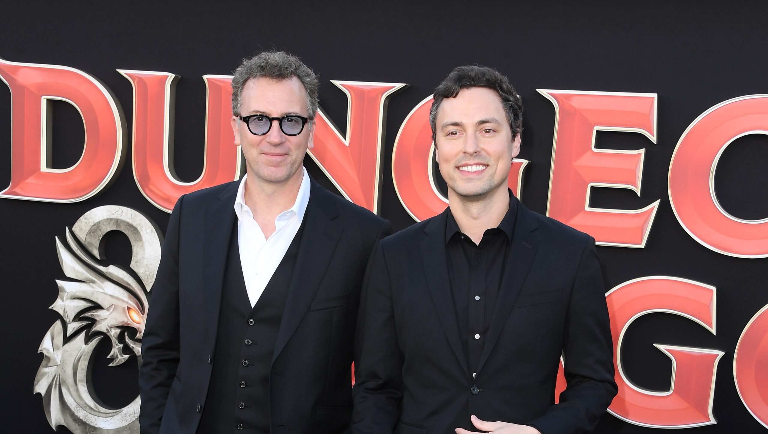 Two men in all black suits posing together on a red carpet.