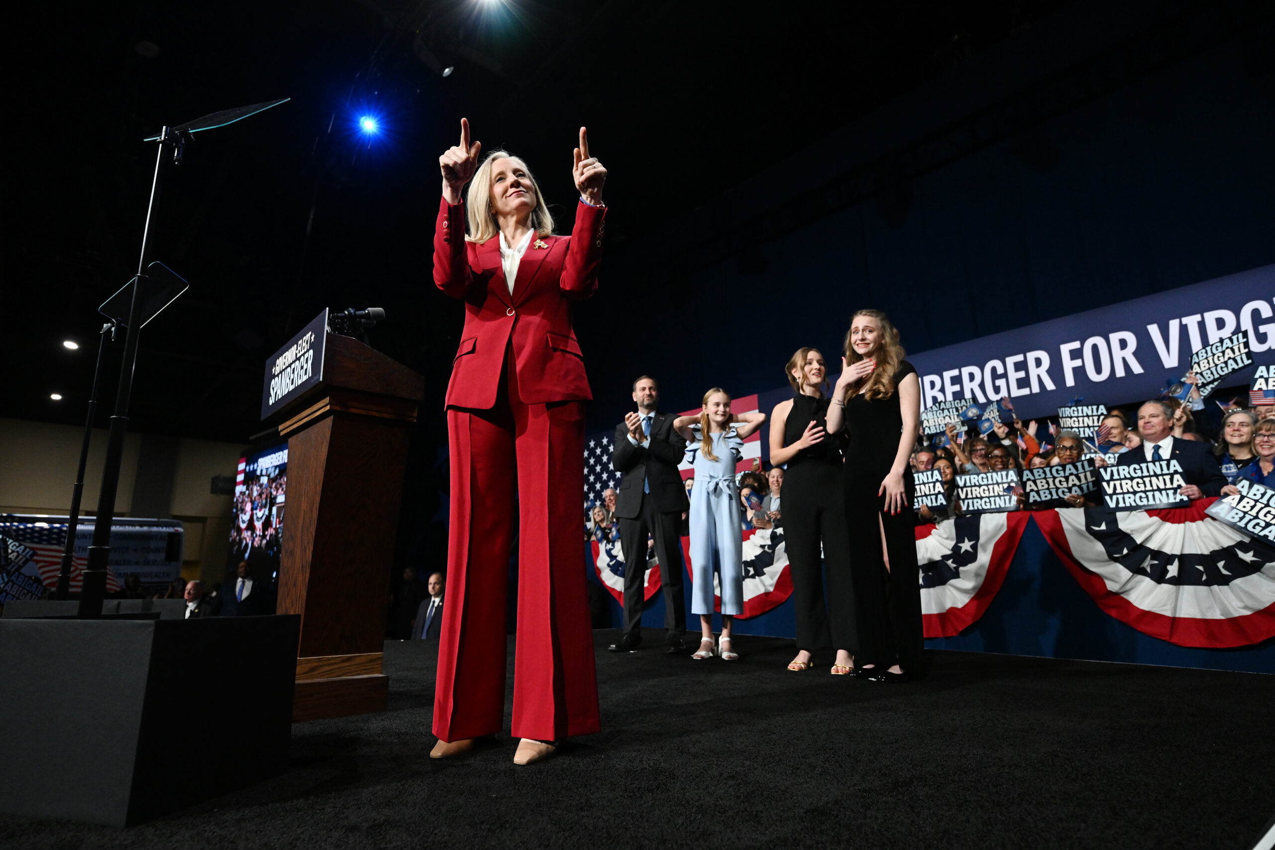 A woman in a red suit stands on a stage and holds up her two index fingers.