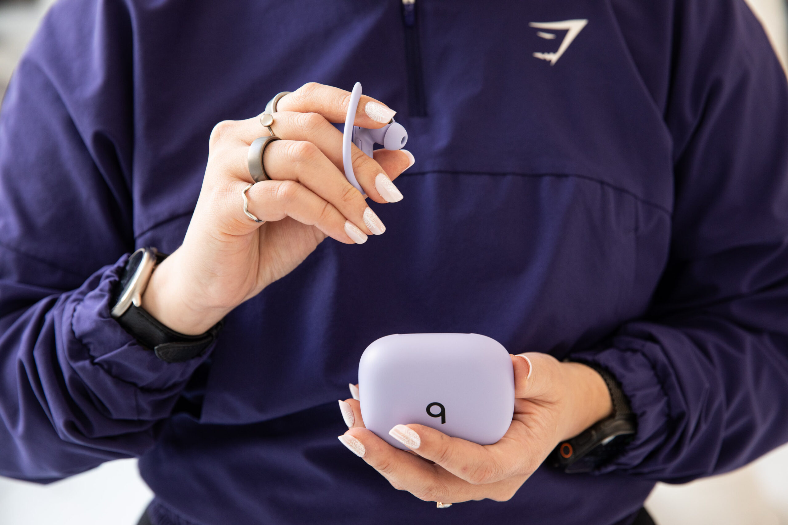 A person wearing a purple workout jacket holding the purple Beats Powerbeats Pro 2 earbuds and case.