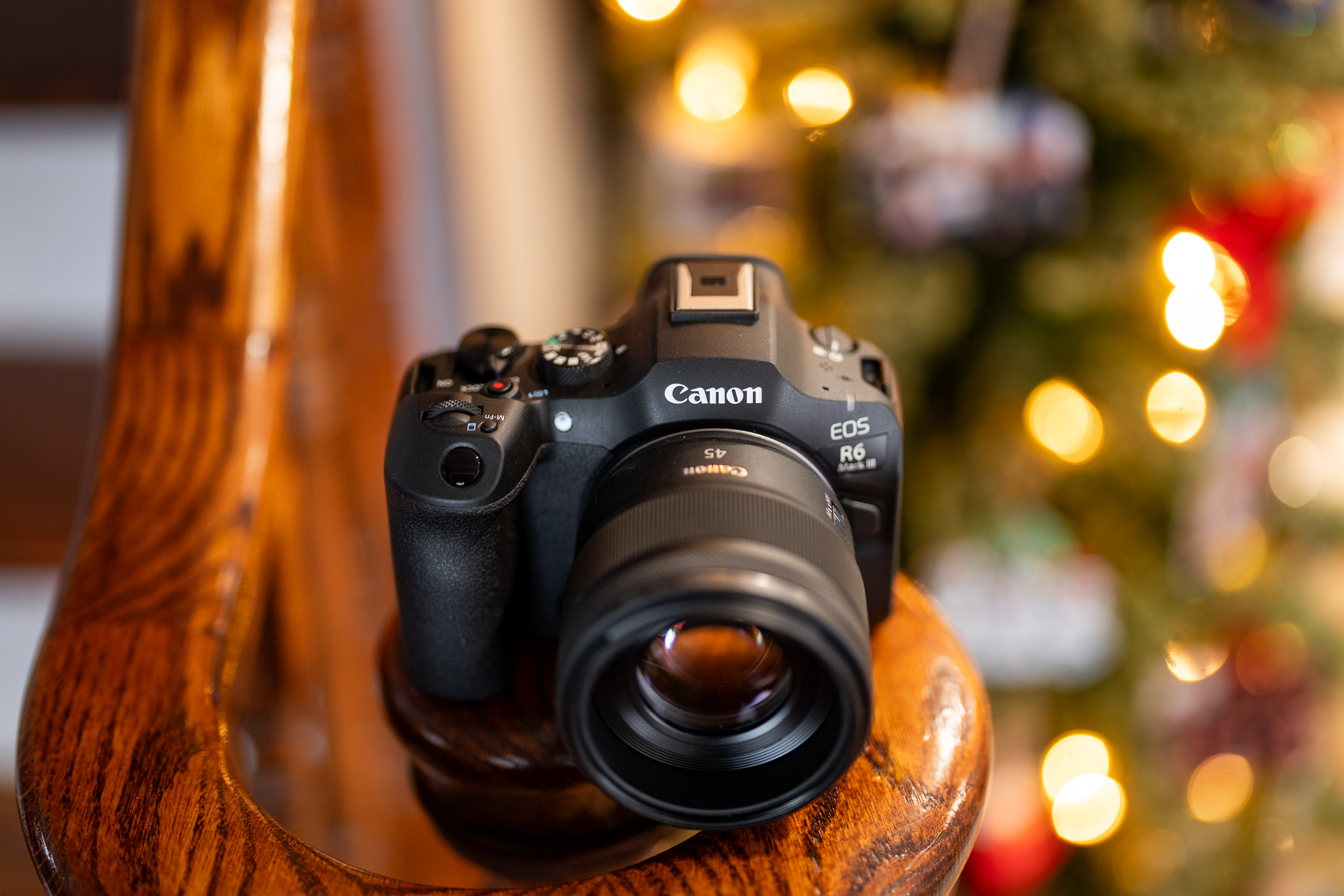 A Canon EOS R6 Mark III camera with RF 45mm f/1.2 STM lens sitting on a wood banister in front of a festive tree and string lights. The background is heavily blurred, with a shallow depth of field.
