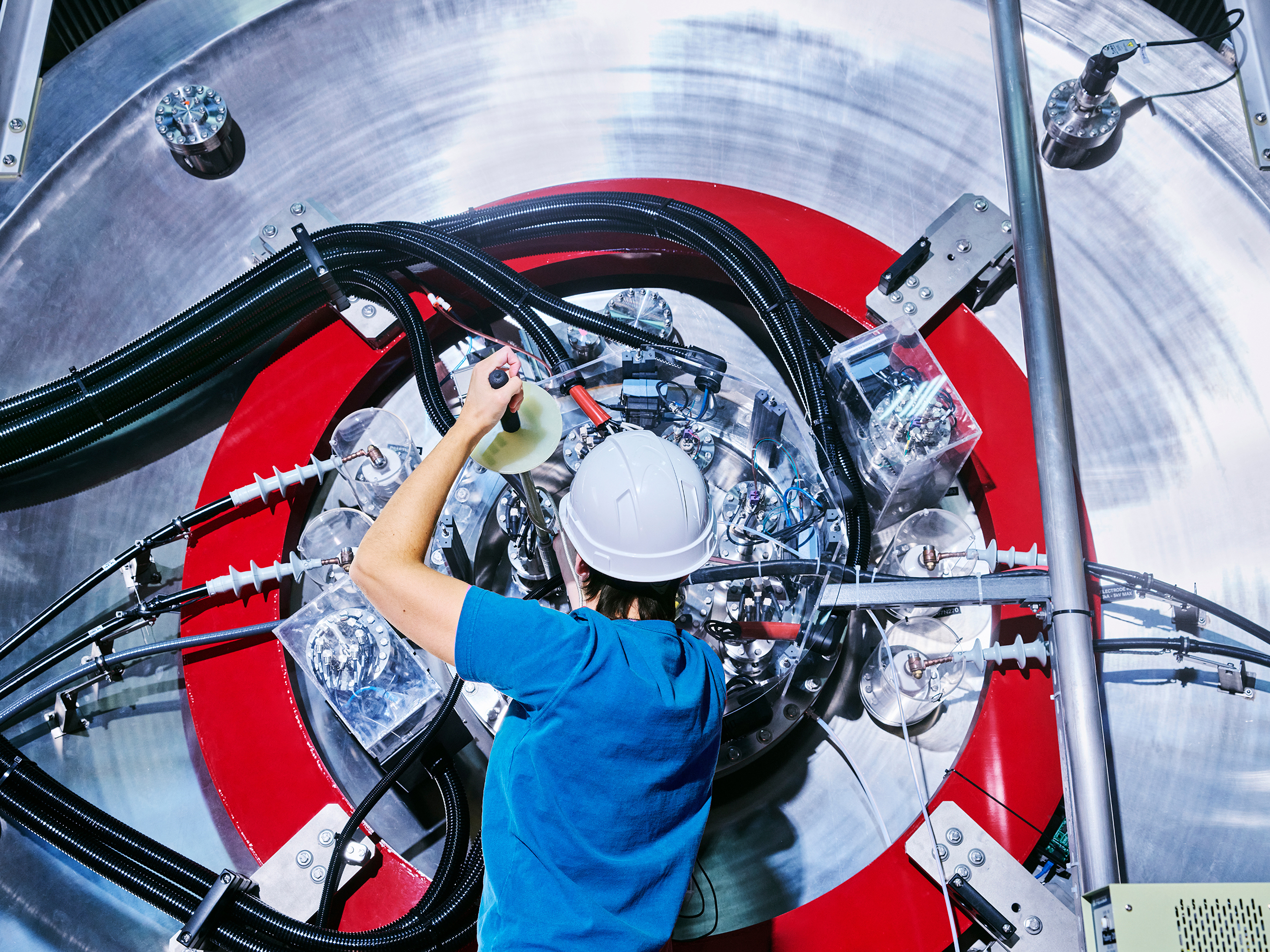 A worker wearing a hard hat works on a large metal machine with a tool. 