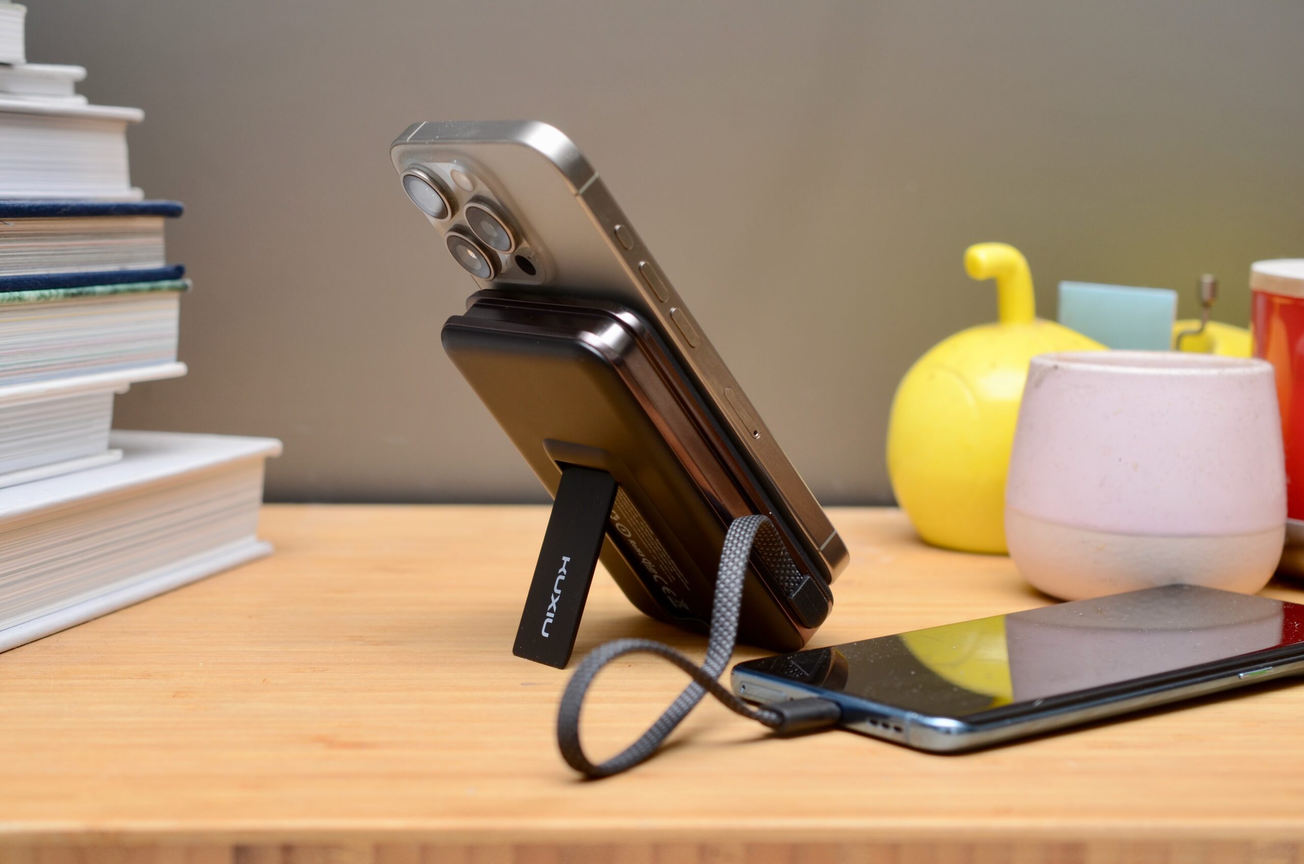 An iPhone rests on the MagSafe connector on the Kuxiu S3 propped up by its kickstand, while the power bank charges another phone resting on a bamboo tabletop where books and trinkets are visible.
