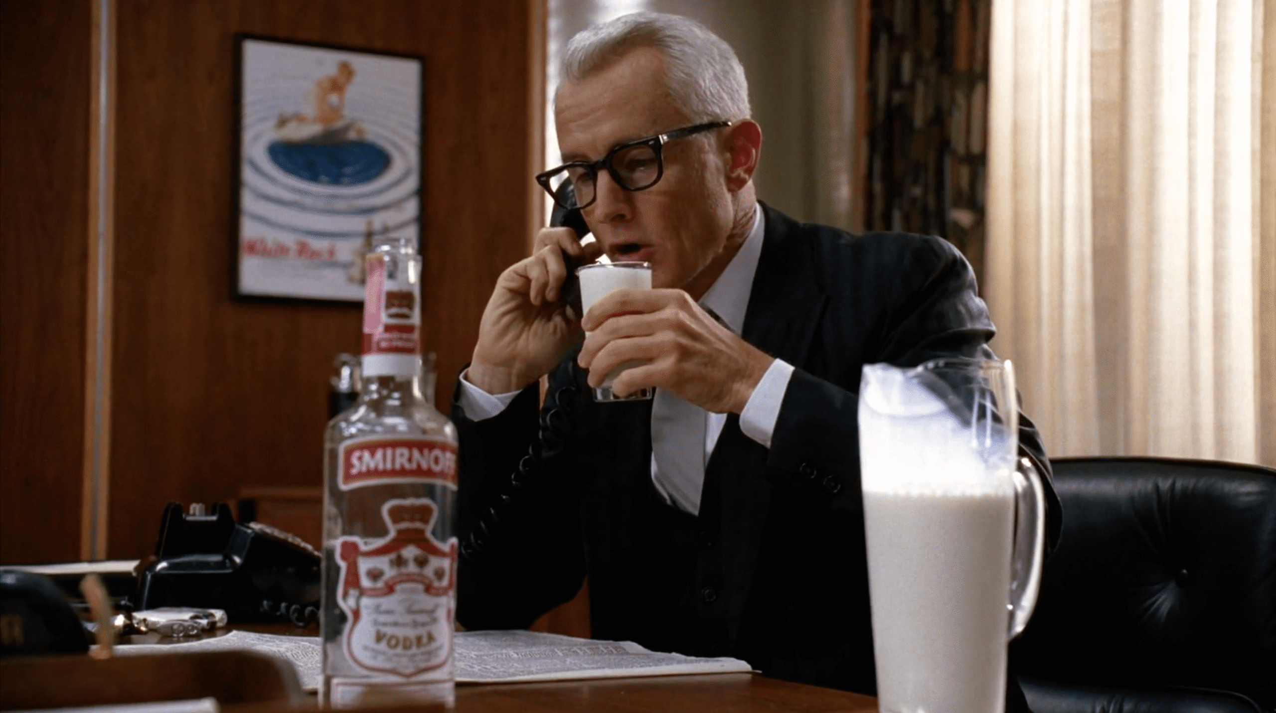 A man in a suit while sitting on a desk and sipping milk from a glass. On the table ie a pitcher of milk and a bottle of vodka.