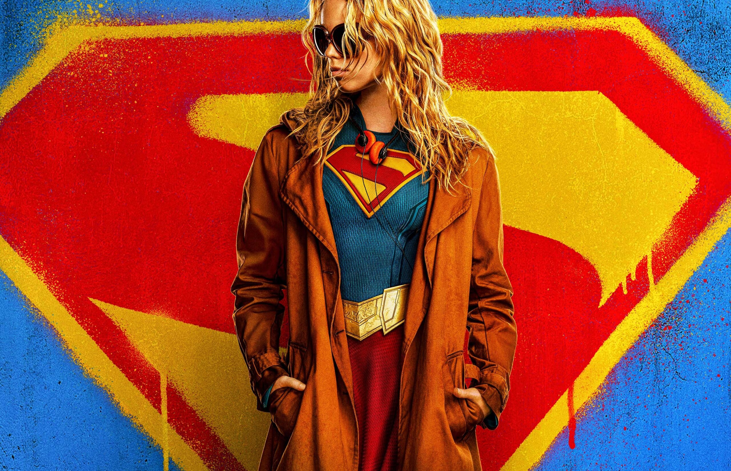 A woman in a blue, formfitting superhero top emblazoned with the Superman logo. The woman is also wearing a red skirt, ret boots, large sunglasses, and a brown trench coat..