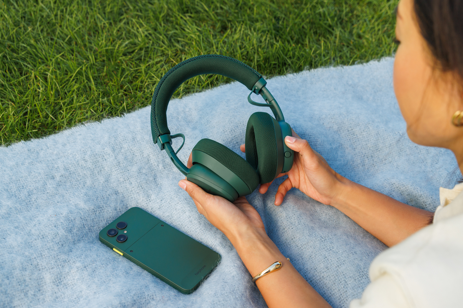 A person lying on a blanket on grass holds the forest green version of Fairphone’s new Fairbuds XL headphones.