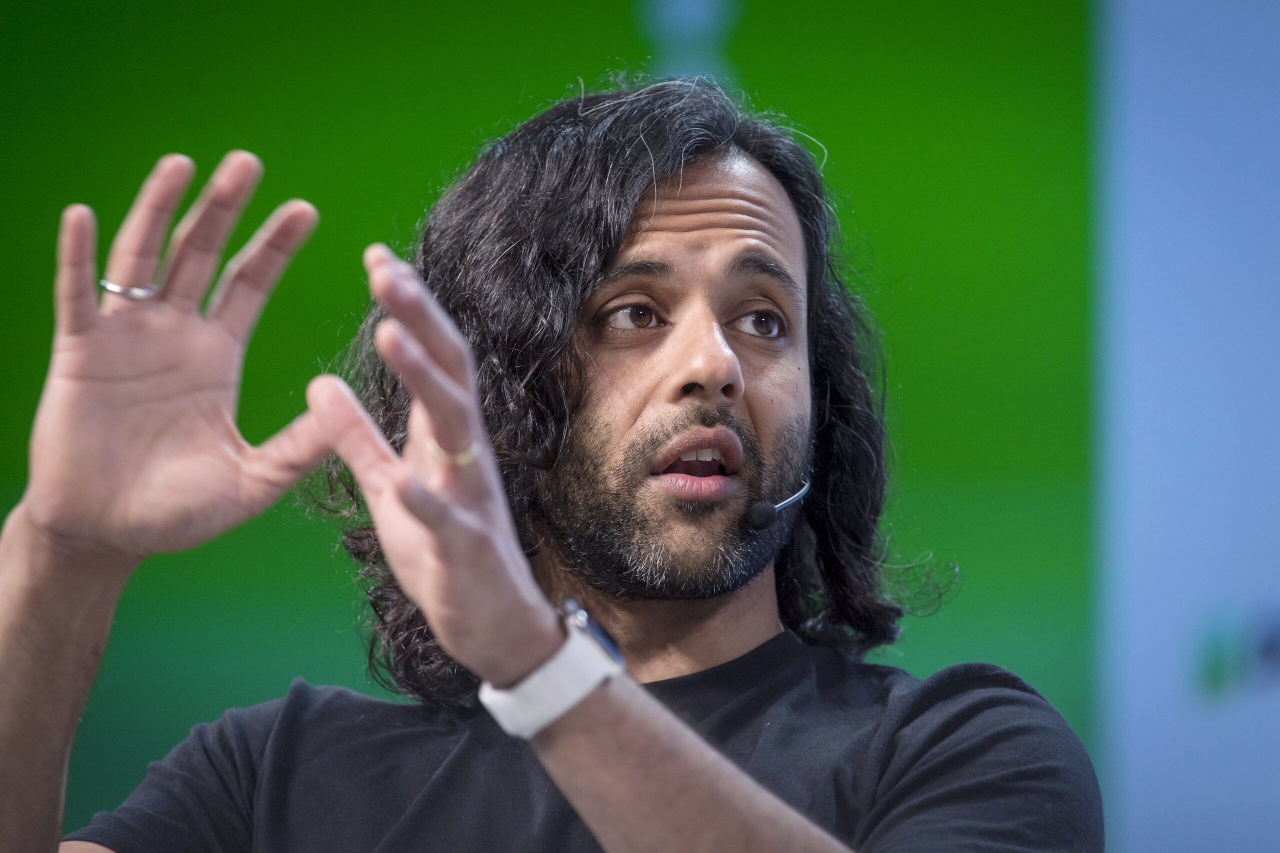 A person with shoulder-length hair speaking with two hands held up in front of their face.