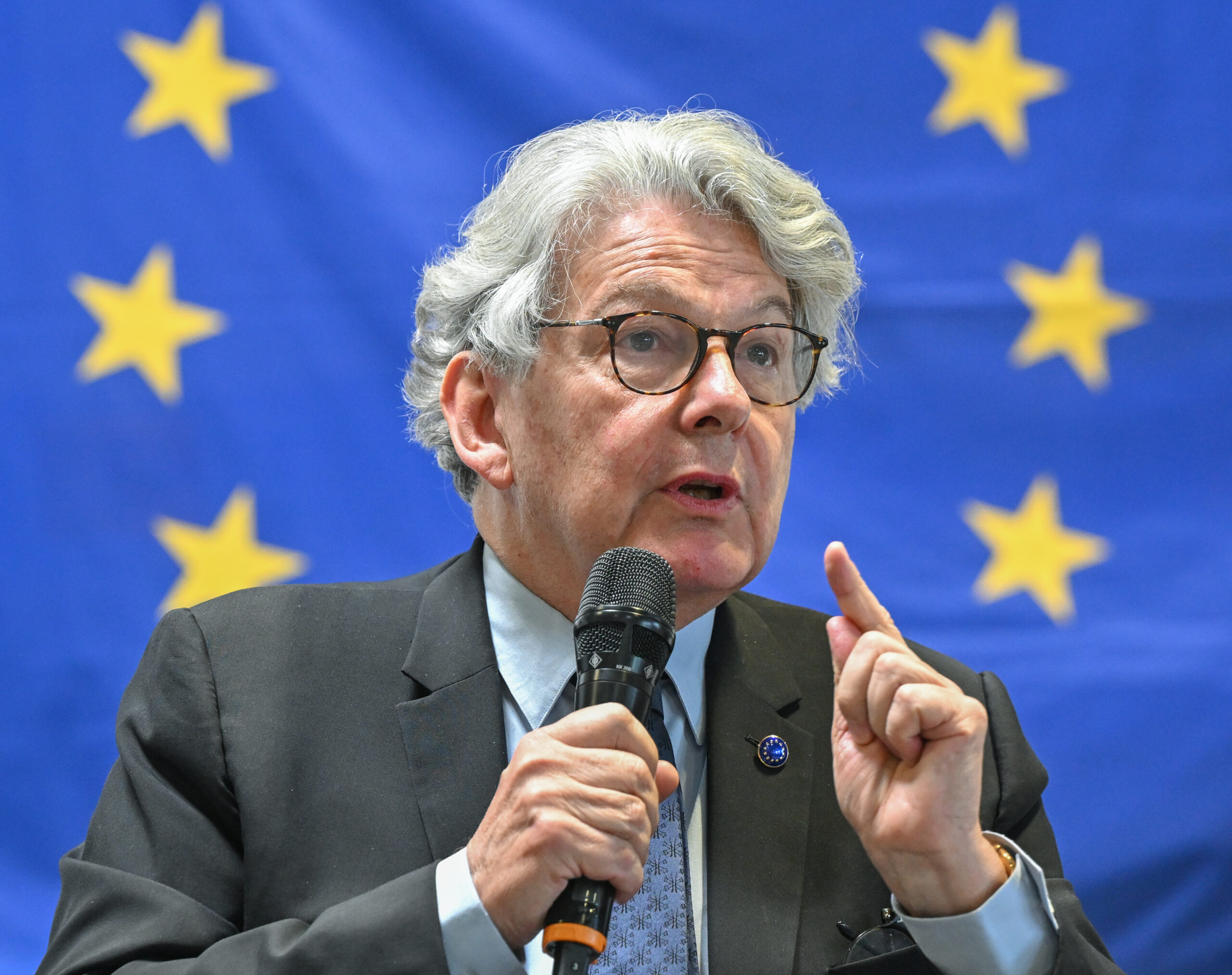 An image of a man in a suit with white hair speaking into a microphone with the EU flag in the background.