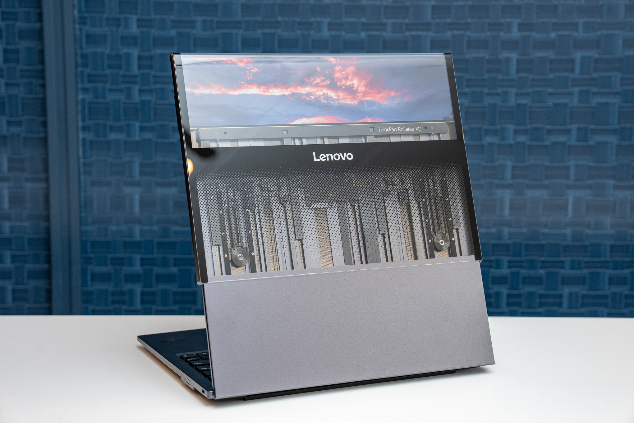 A Lenovo ThinkPad Rollable XD concept laptop with its lid open and rollable display extended, viewed from the back. Its flexible OLED screen wraps around to the outer part of its lid, which is partially seethrough to show the motors and pulleys inside.