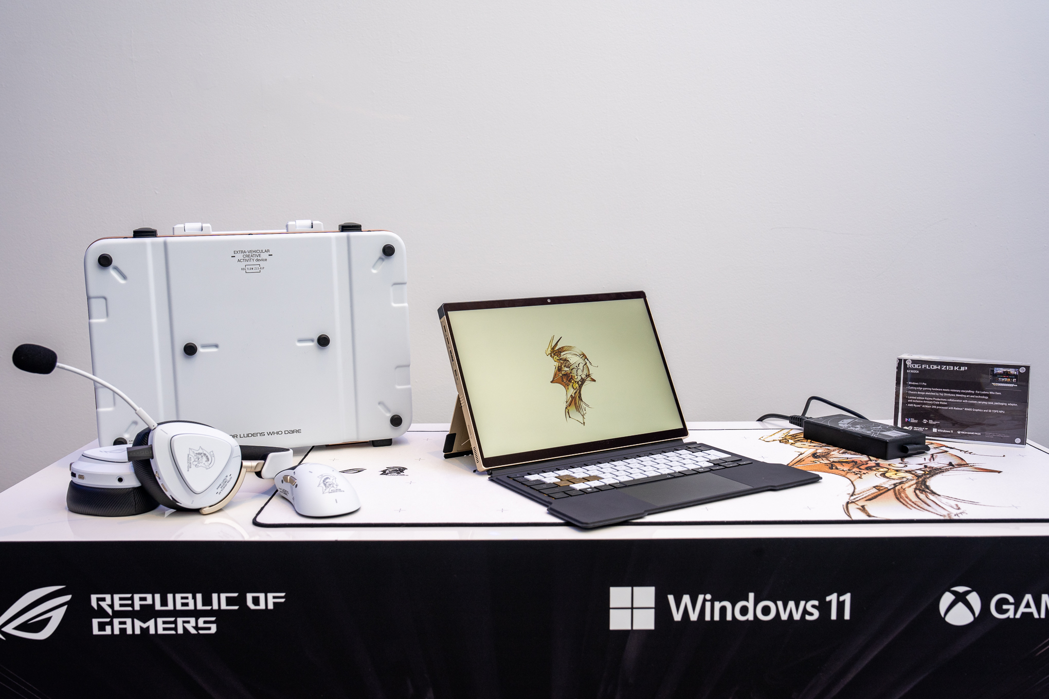 The Asus ROG Flow Z13 x Kojima Productions limited edition tablet on a white table beside its various accessories: a desk mat, headset, mouse, attache case, keyboard cover, and power adapter.