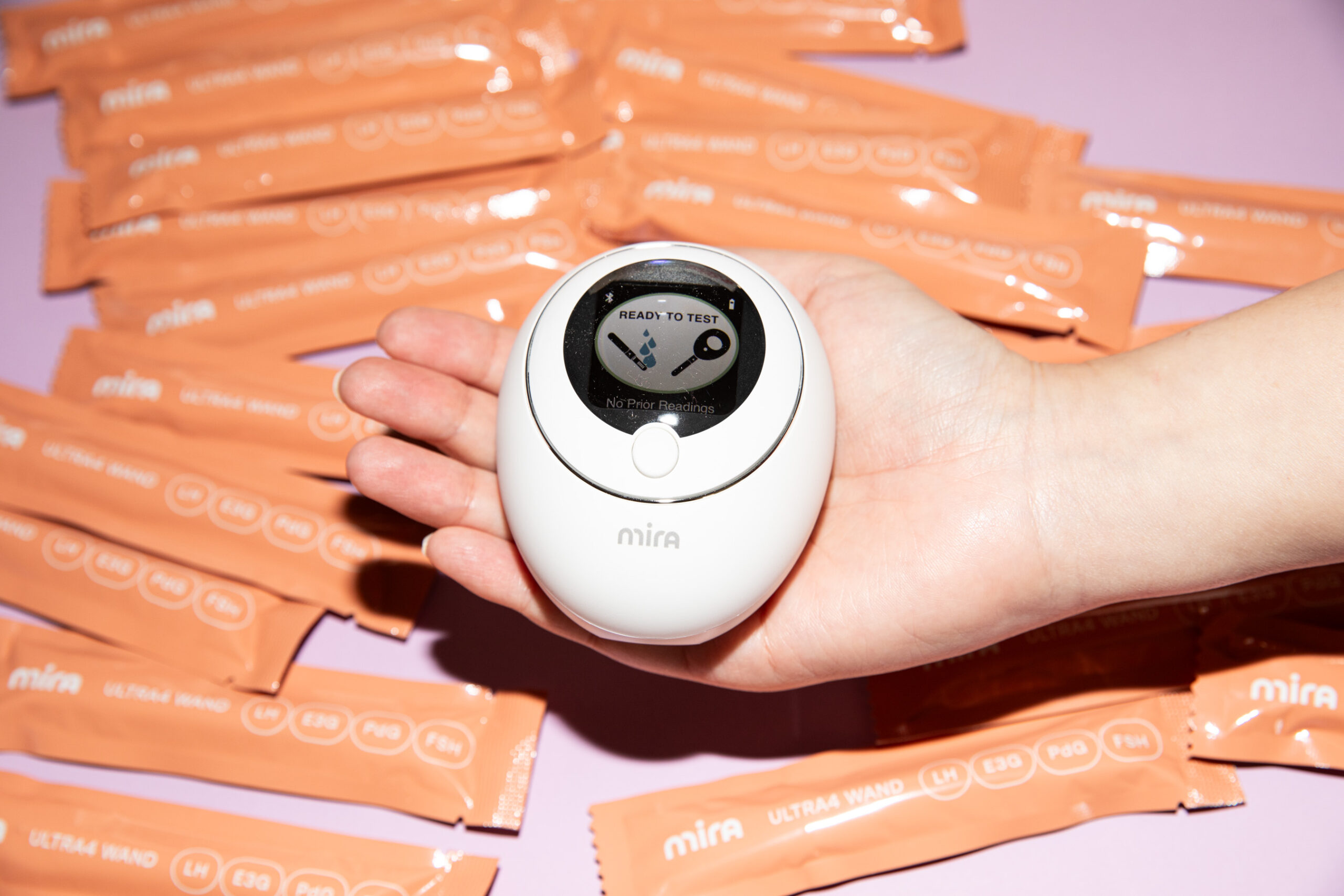 Top-down view of the Mira Analyzer in someone’s hand. It’s egg-shaped with a screen in the middle. In the background are Mira Ultra4 Wands scattered about in coral packaging on top of a lavender backdrop.