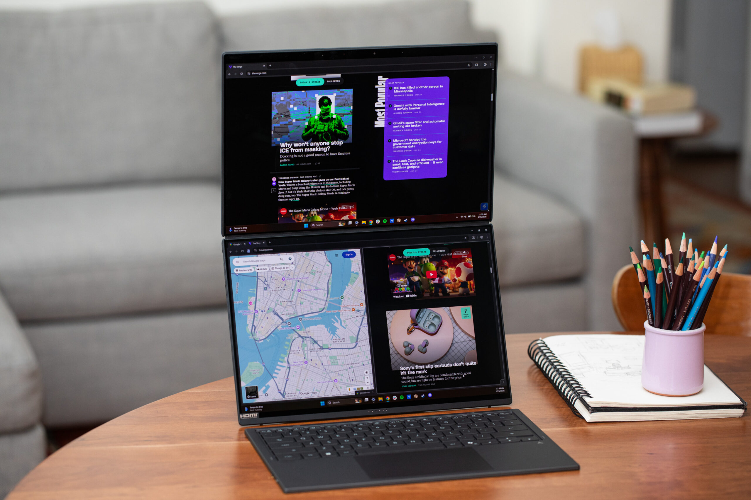 An Asus Zenbook Duo (2026) dual-screen laptop sitting on a desk beside a notepad and cup of colored pencils. It is propped up by its built-in kickstand, displaying both its screens with content on each. Its keyboard and trackpad are detached and placed in front for wireless use.