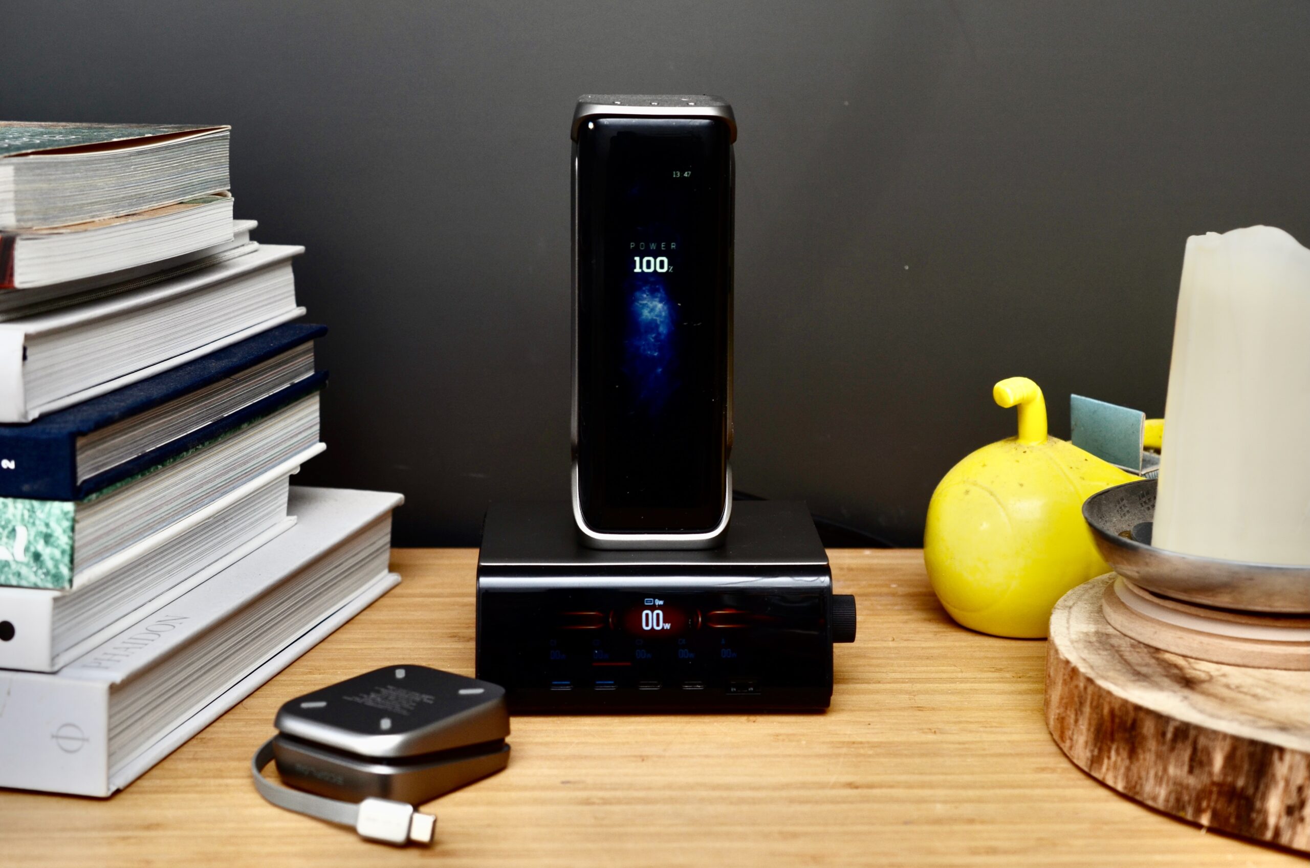 A power bank sits on top of a dock, both with large display, while a retractable cable module sits on a table surrounded by books in the foreground.