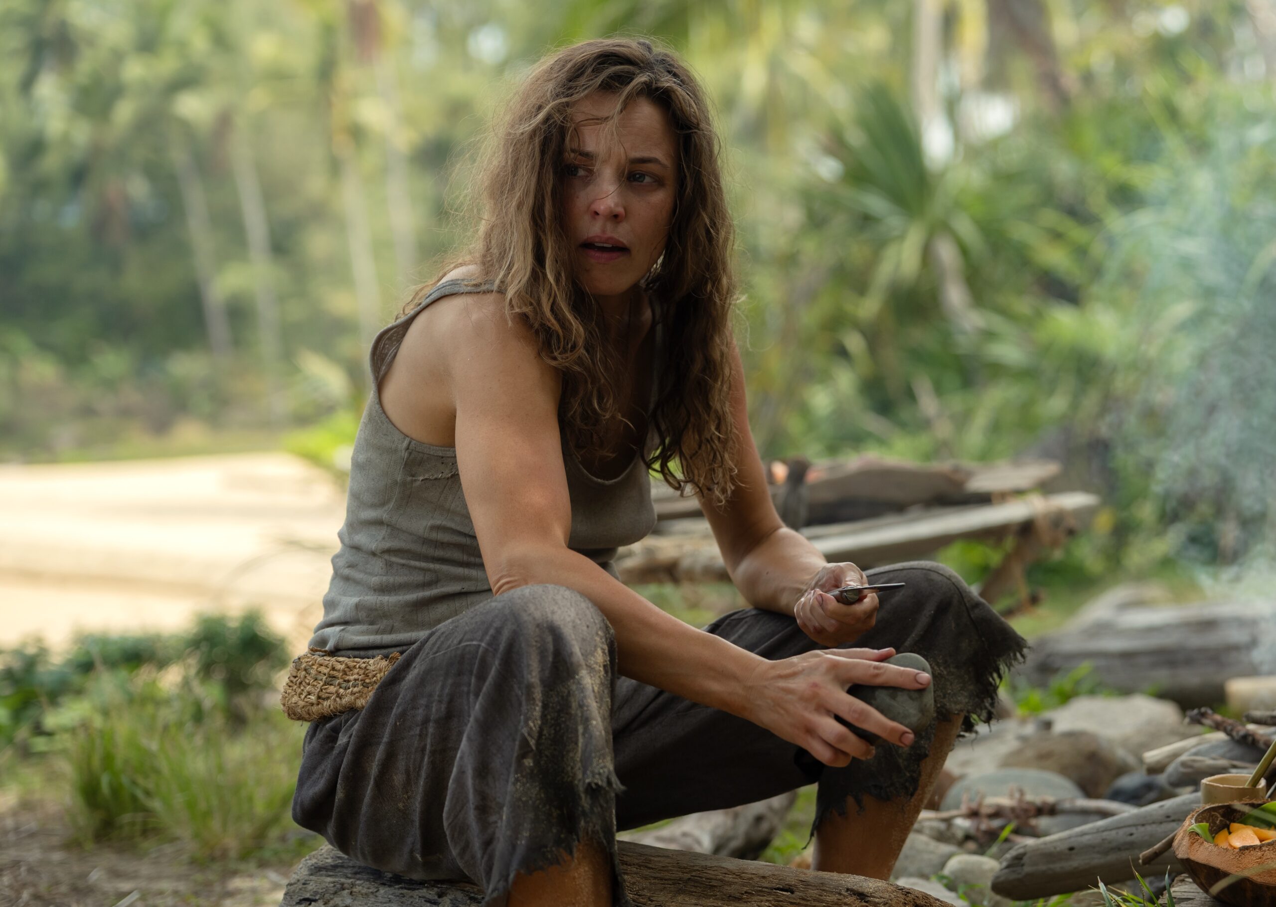 A woman sitting by a fire on a beach. In front of the woman is a basket woven out of leaves.
