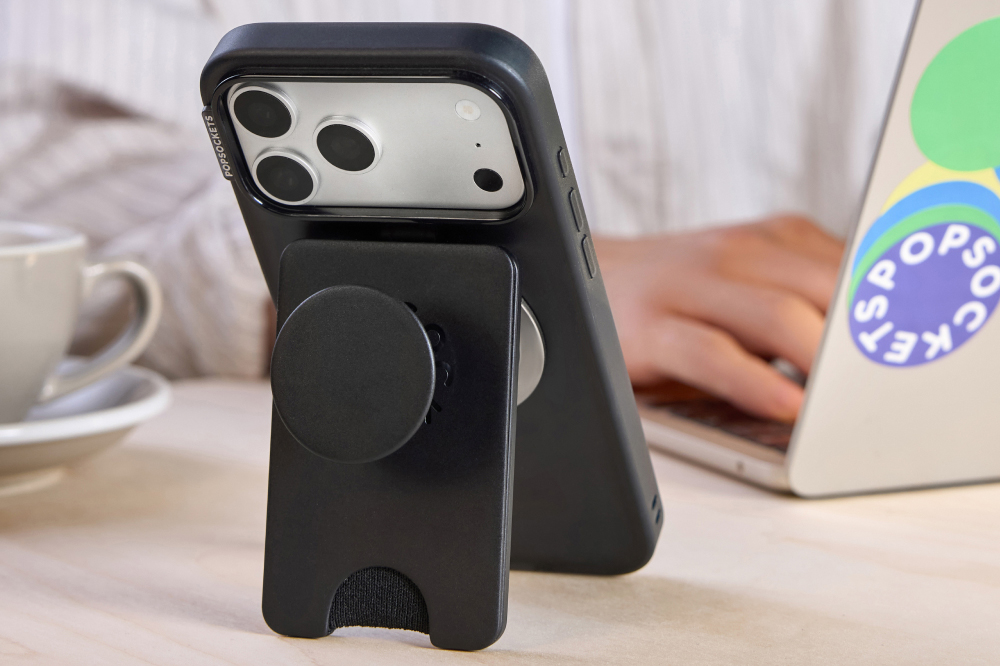 The black version of the PopSockets Kick-Out PopWallet attached to an iPhone on a table next to a person working on a laptop.
