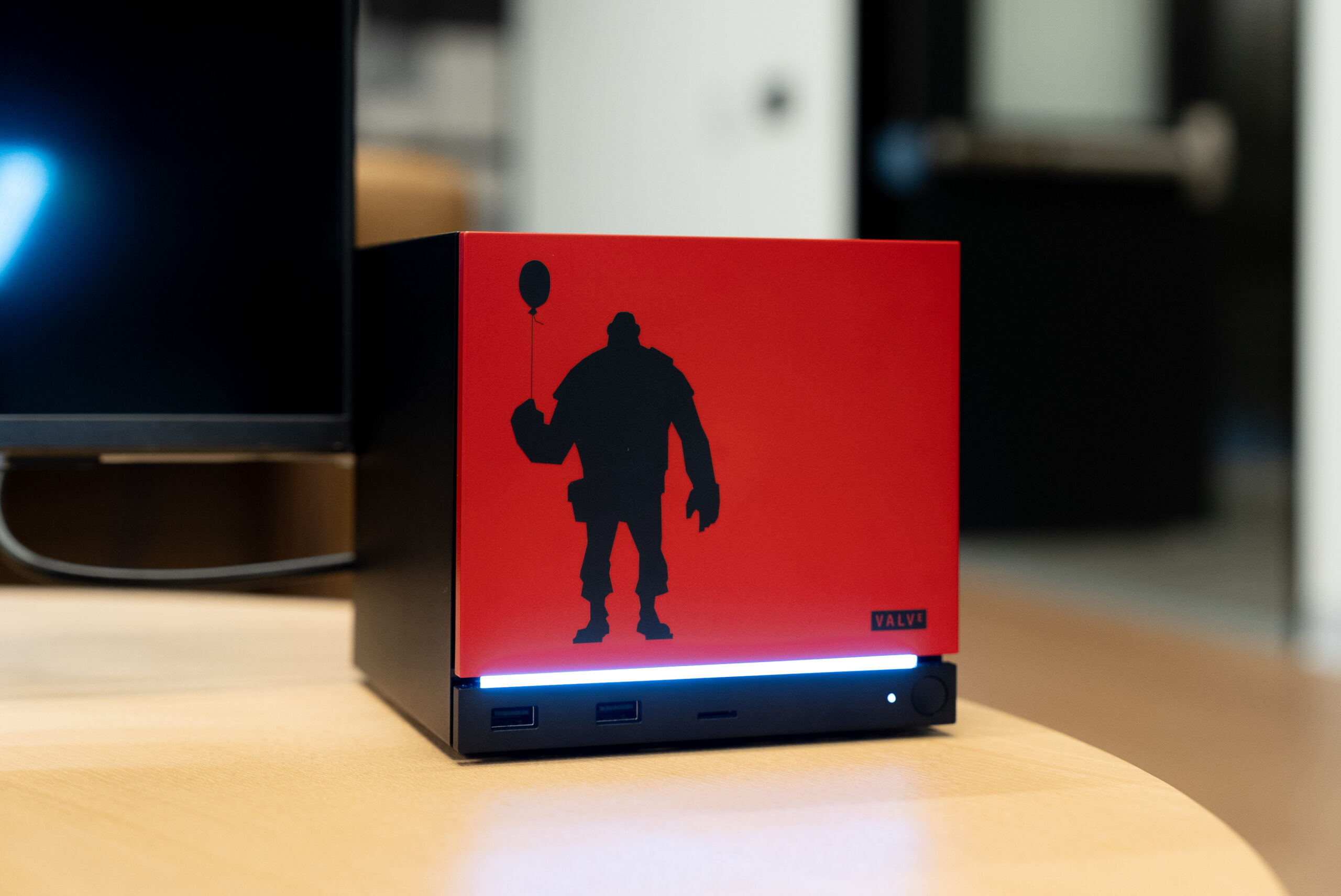 A small black cube with USB ports on the bottom of the front panel, a thin glowing light bar above that, and the remaining seven-eighths of the front panel red with a Team Fortress character in silhouette.