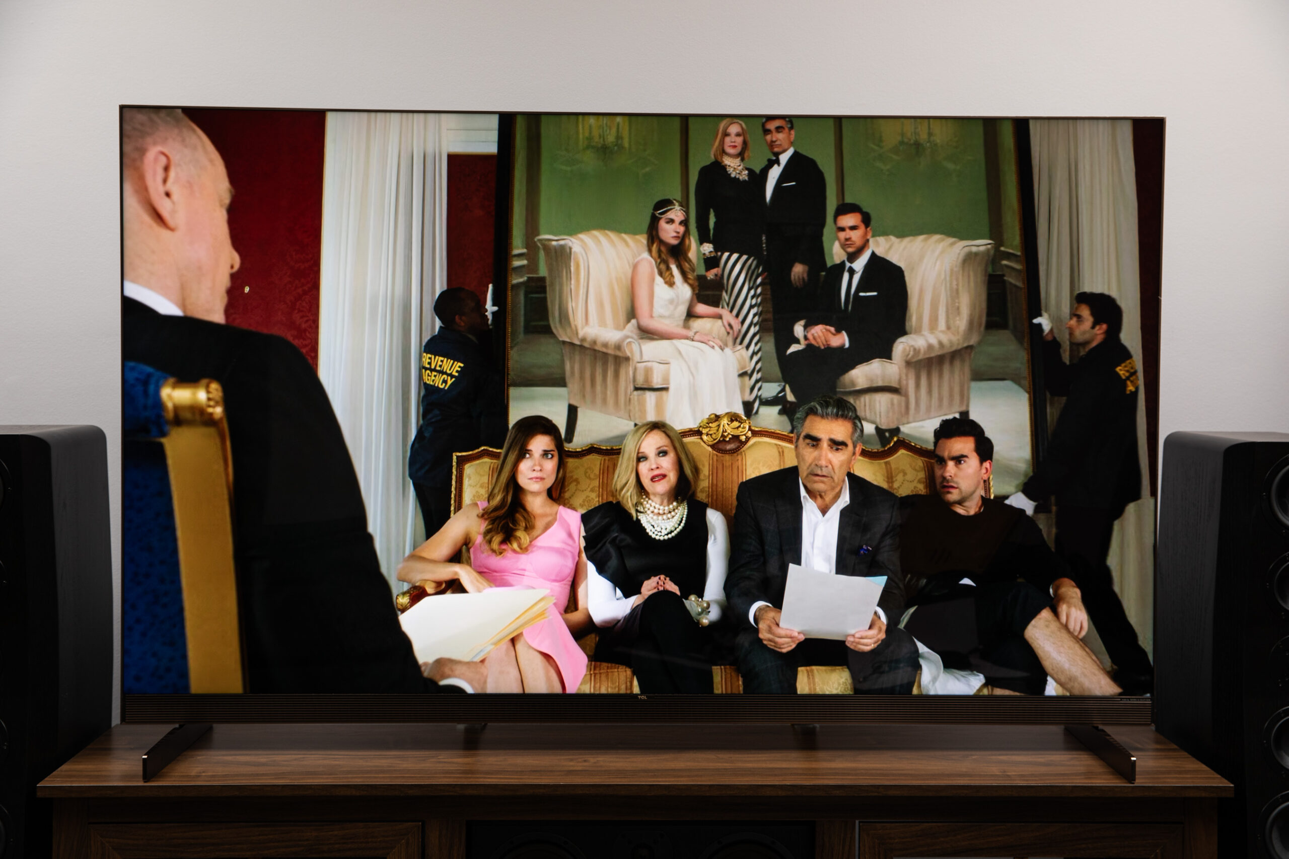 The TCL X11L SQD-Mini LED TV displaying an image from Schitt’s Creek on a dark wood TV stand with SVS speakers on either side.