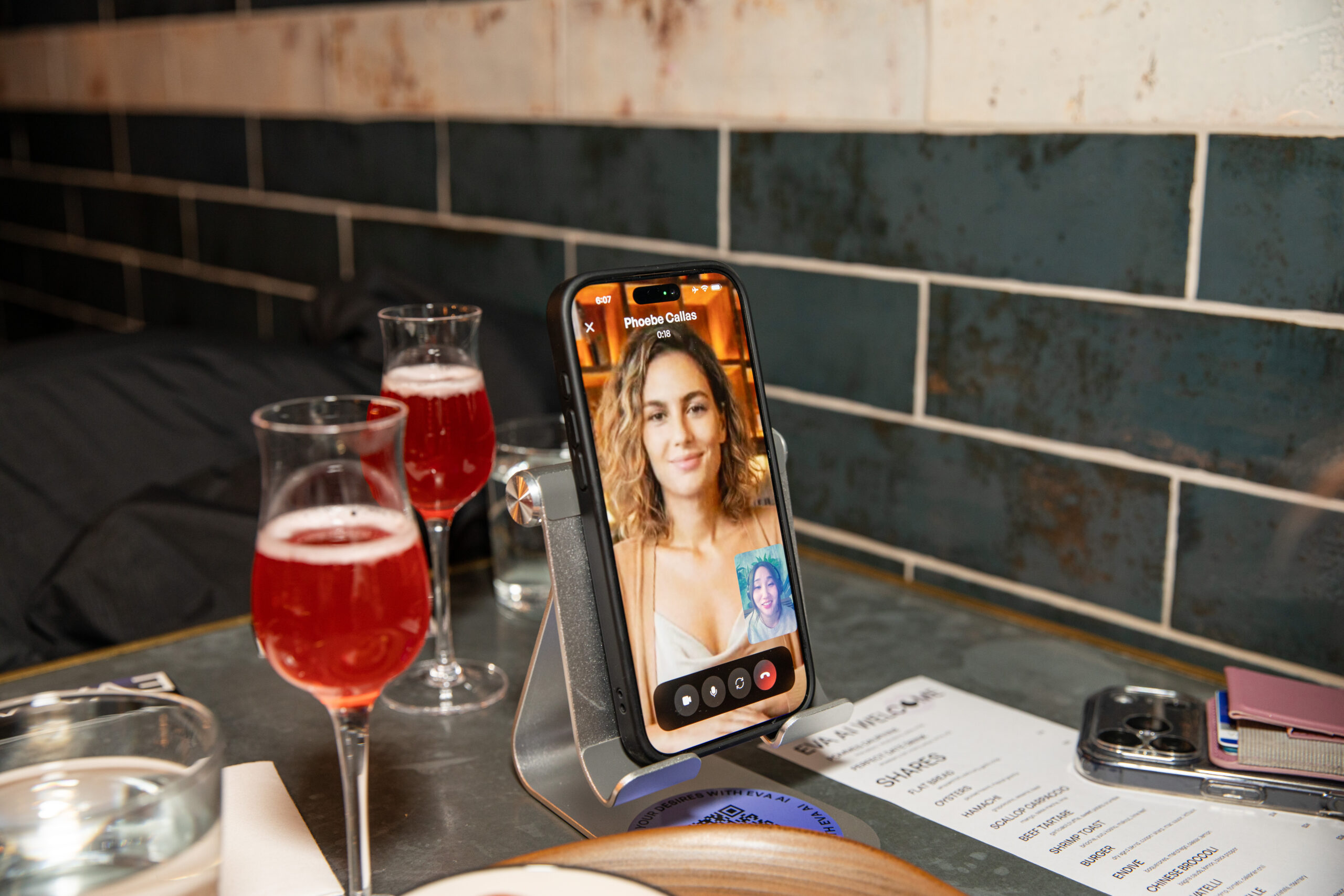 A phone showing an AI companion named Phoebe Callas with a menu, two glasses of a red nonalcoholic drink and a menu are shown.