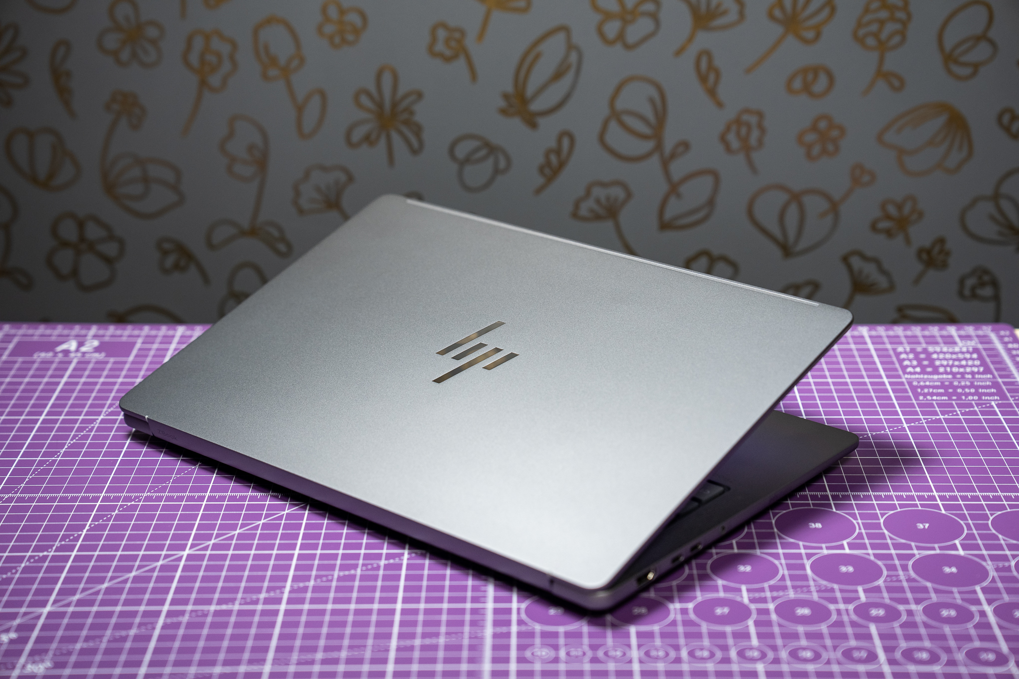 An HP ZBook Ultra G1a laptop mostly closed and showcasing its lid logo. It’s sitting on a purple cut mat in front of a gray wall with a gold floral design.
