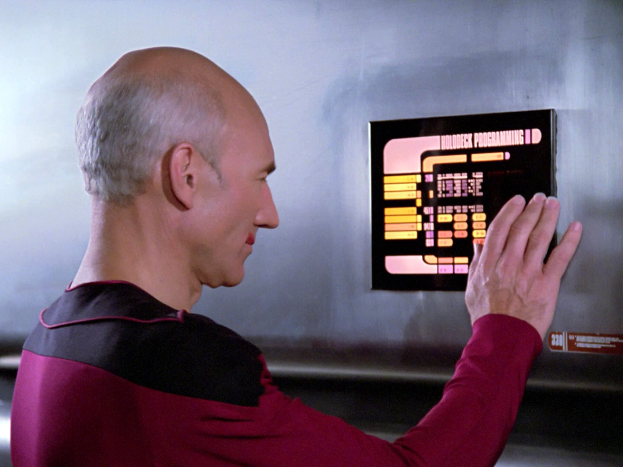 a man in a futuristic uniform looks at a wall-bound tablet with colorful buttons