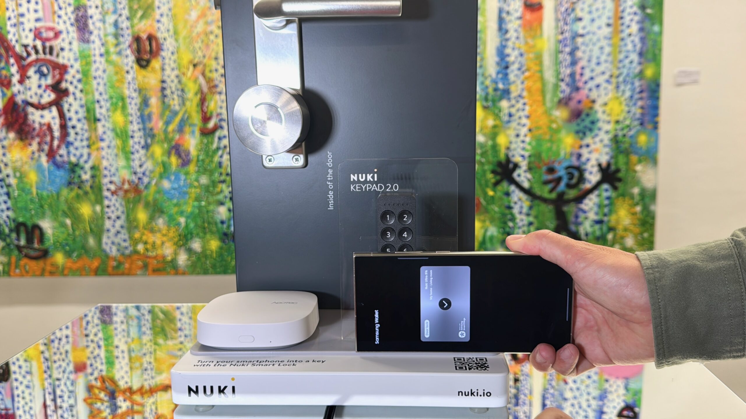A hand holding a smartphone in front of a mockup of a door. There’s a smart lock in the door mockup and the phone is displaying a lock.