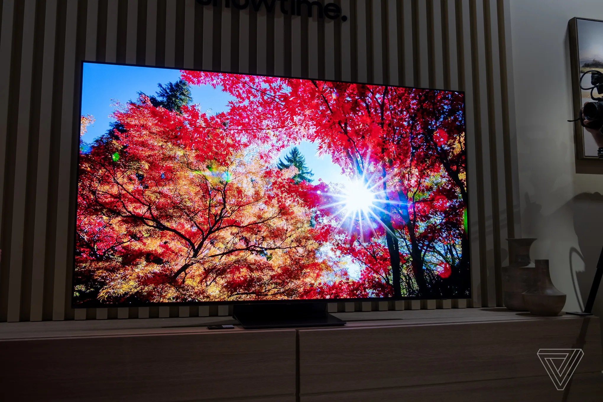 In a dark room, against a backdrop of wood panel slats, a brilliant TV screen with almost no bezel displays sun streaming through red and yellow fall color leaves.