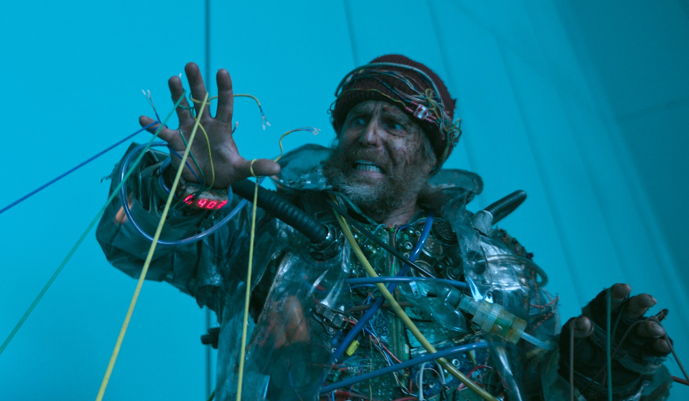 A man in a suit made out of old pieces of junk technology. The man is looking in fear at his hand, which is being pulled apart by a series of electronic wires.