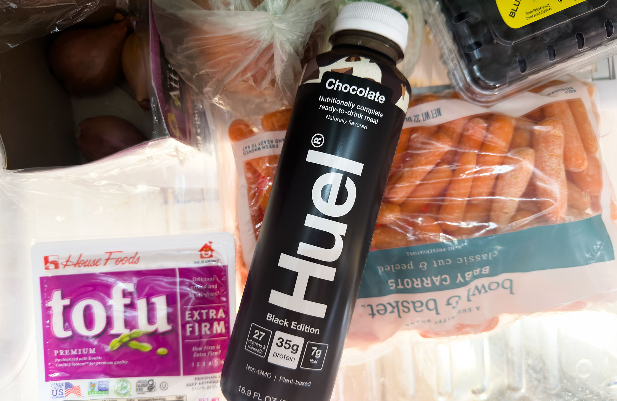 A bottle of Huel ready-to-drink in the chocolate flavor in a refrigerator door surrounded by blueberries, carrots, tofu, and shallots