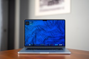A 15-inch MacBook Air on a table with its screen open and on. It’s running macOS Tahoe 26 and showing the desktop with a blue wallpaper.