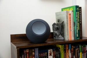 The 2025 Amazon Echo Studio sitting on a wooden shelf next to a collection of books, with a white wall behind it.