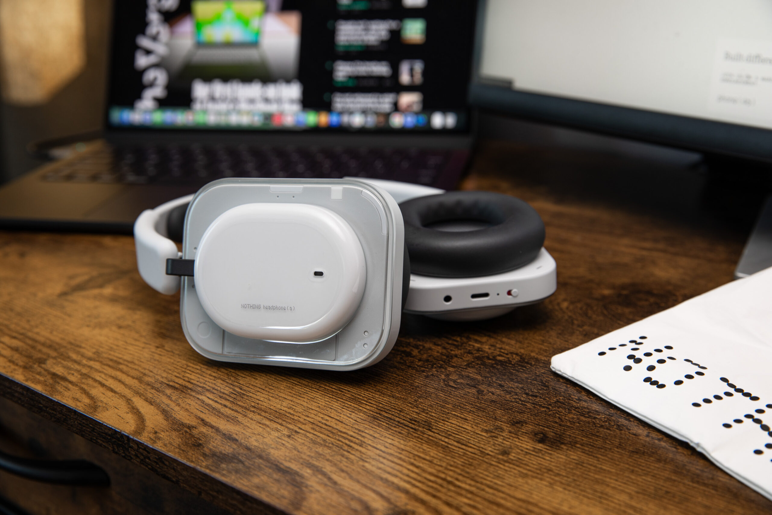 The Nothing Headphone A in white on a wooden desk with the carry bag off to the side and an open laptop behind.