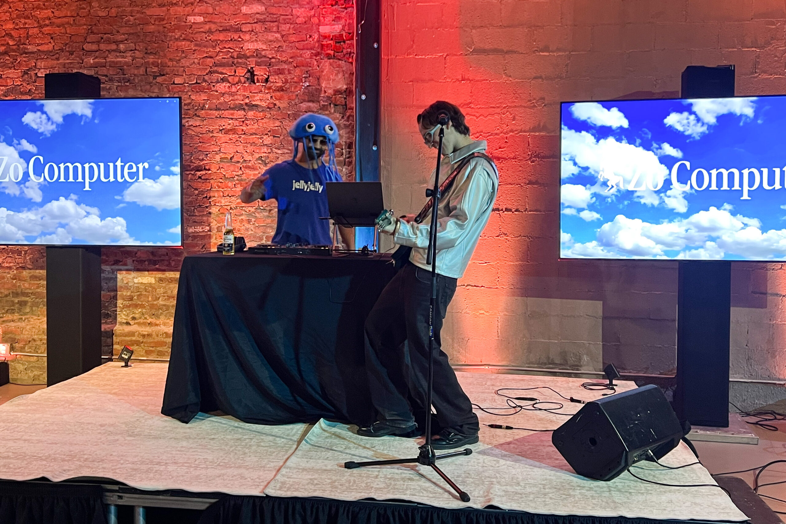 A guitarist and a DJ in a blue plush jellyfish hat jam on a small stage.