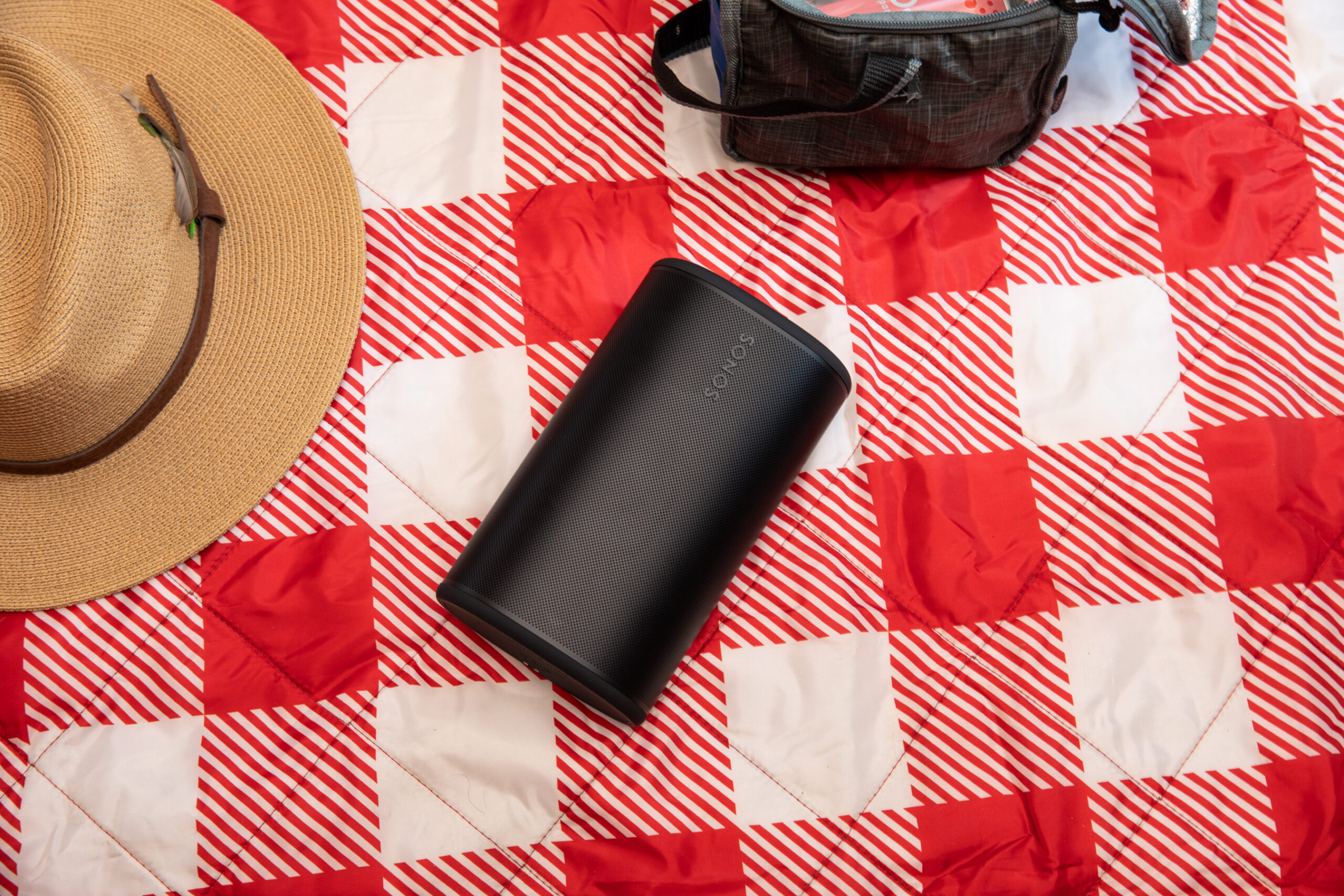 The Sonos Play on a red checkered blanket next to a soft-sided cooler and a sun hat.