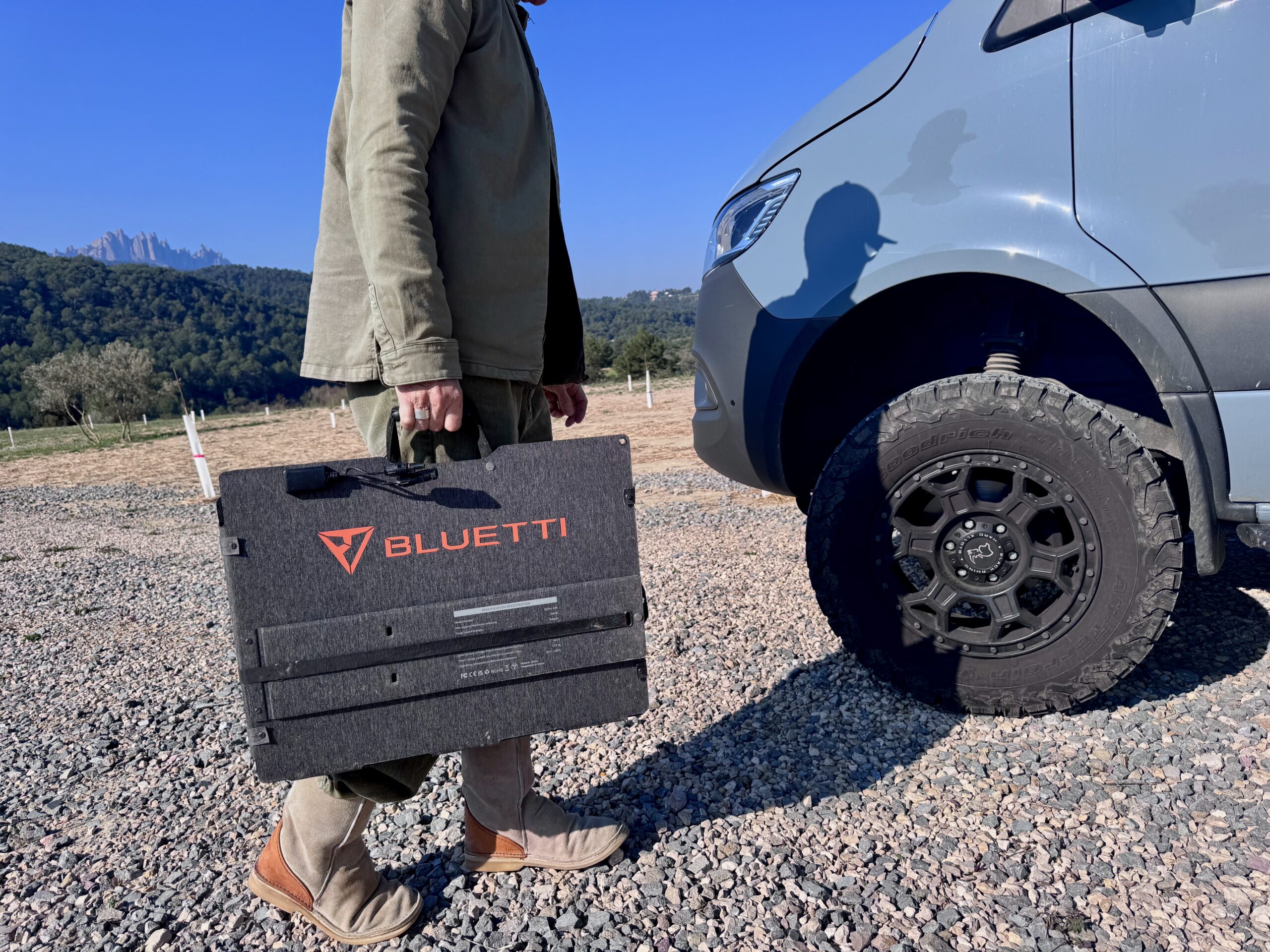 Carrying the Bluetti Sora 500 panel by its handle with the wheel of a van and mountain range in the distance.