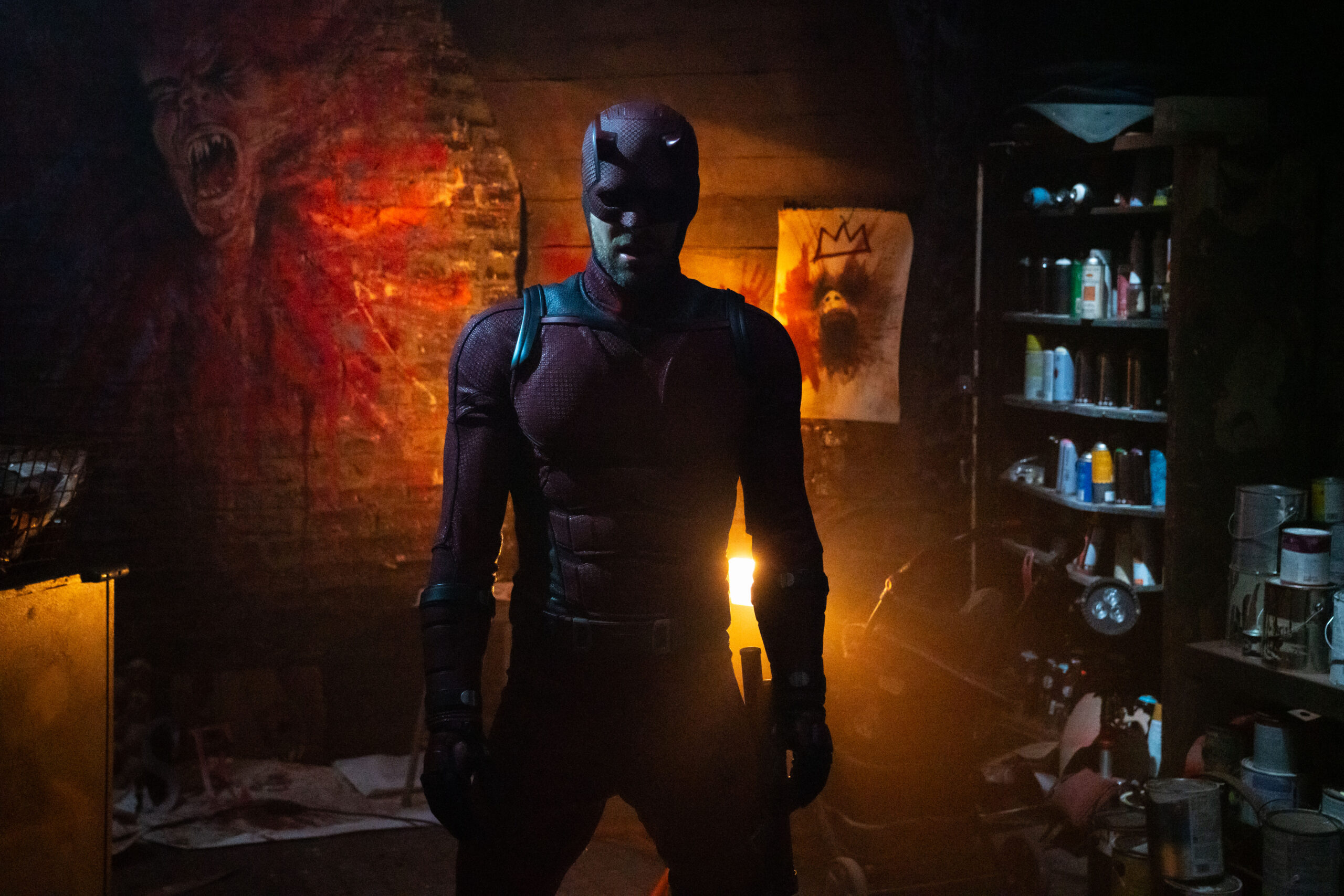 A man wearing a dark red full-body superhero costume and standing in a dark room.