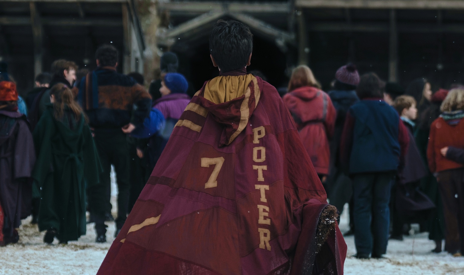 A small boy in a red clock that has the number seven and the name “Potter” emblazoned on it in yellow. The boy has his back turned to the camera as he walks towards a group of people in winter clothing.