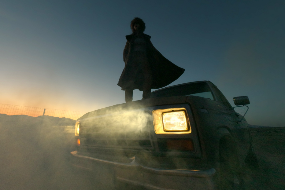A person standing on the hood of a pickup truck with its headlines shining through smoke.