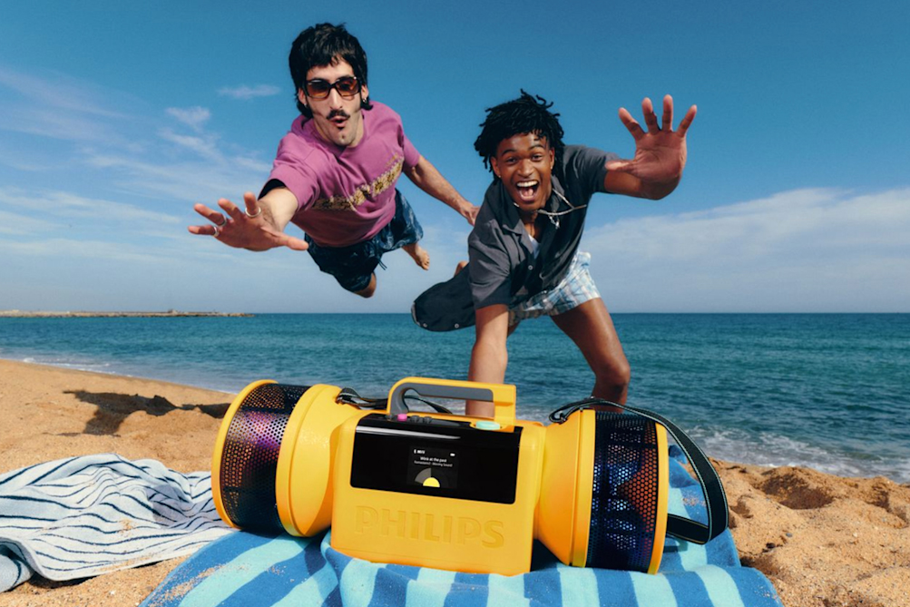 Two people on a beach dive towards a Philips wireless speaker.