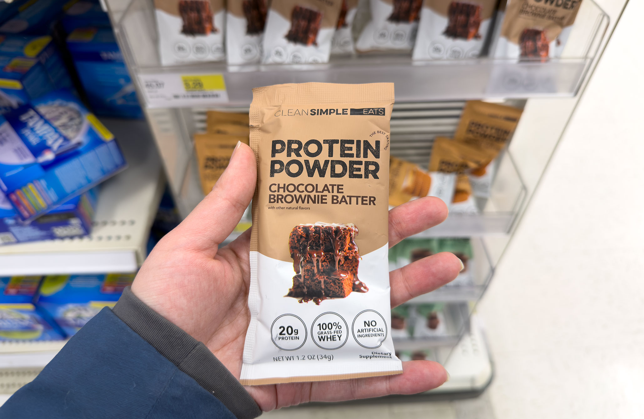 Top-down closeup of a hand holding a protein powder packet in a chocolate brownie batter flavor in the tampon aisle of a Target.