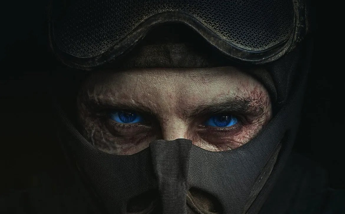A tight shot of a man with striking blue eyes. The man’s face is almost entirely covered by a gray mask.