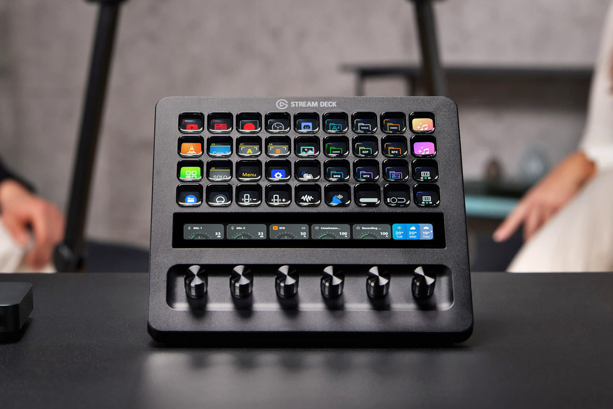 The Stream Deck Plus XL sitting on a desk in front of two people in the background.