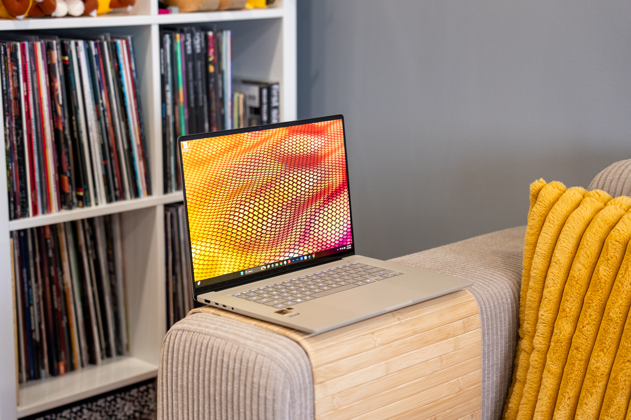 A beige Asus Zenbook A16 laptop on a beige couch in front of a shelf of vinyl records.