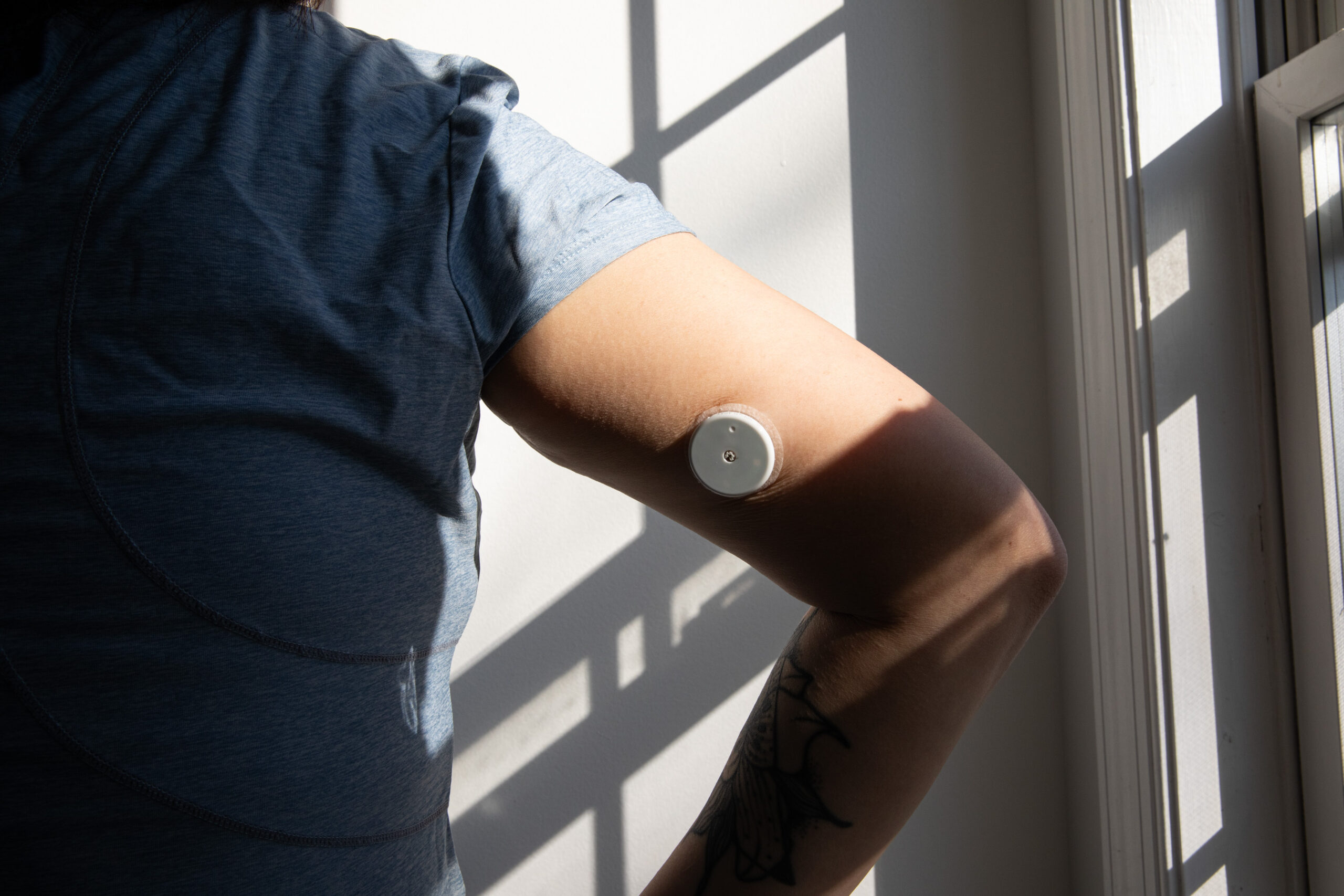 Back view of senior reviewer Victoria Song’s arm while wearing the Abbott Lingo CGM. There are shadows from a window